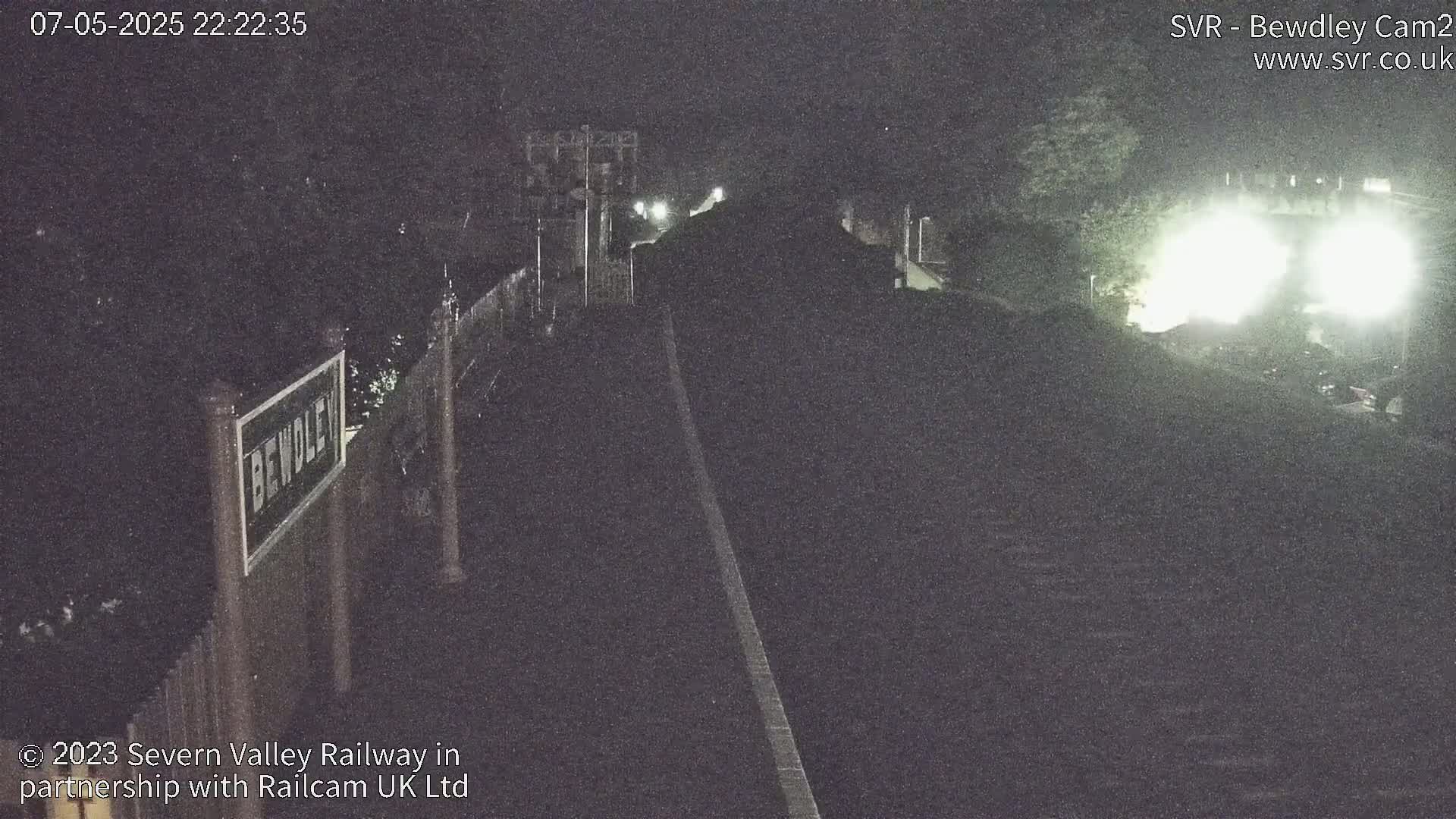 A dimly lit nighttime train scene shows a train passing by a station platform under dark skies.