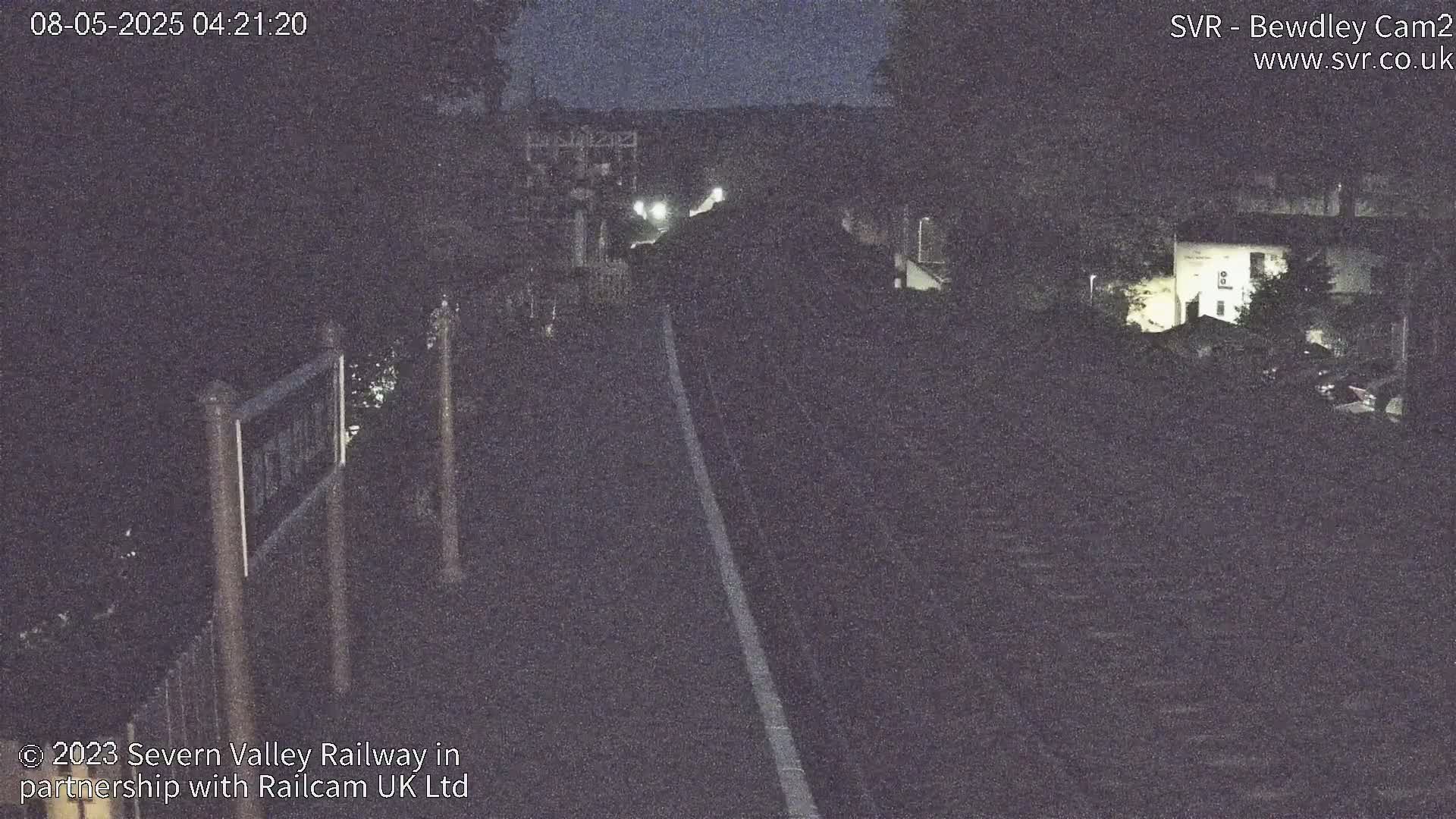 A nighttime view of a railway track, dimly lit, with a signpost visible on the left and buildings in the distance.
