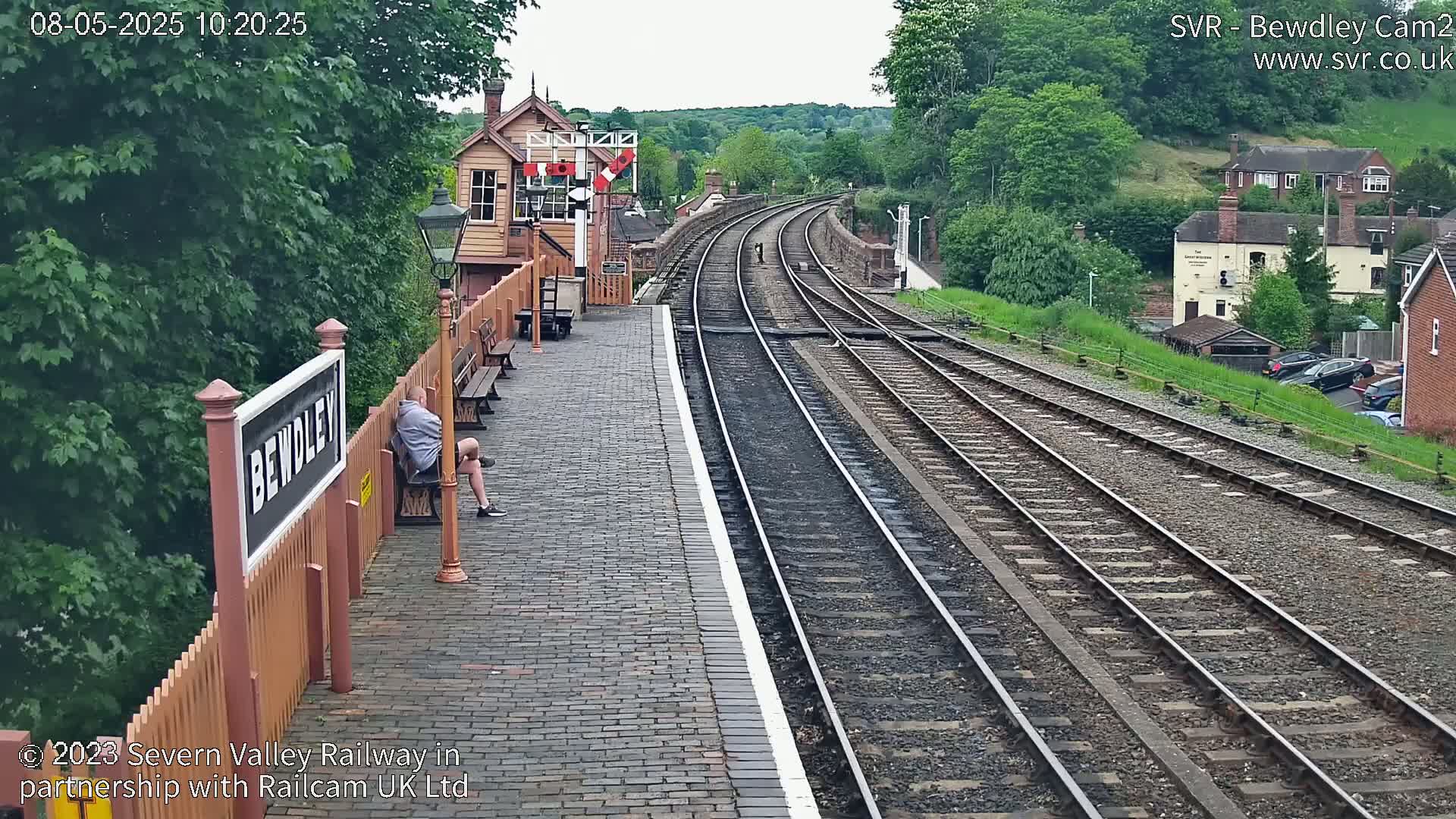 A person sits on a bench at a train station on a partly cloudy day, overlooking several sets of train tracks curving into the distance.
