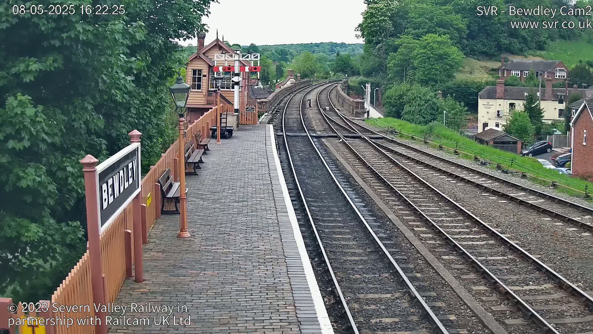 A train station platform with multiple tracks extends into the distance under an overcast sky, surrounded by lush green trees and buildings.