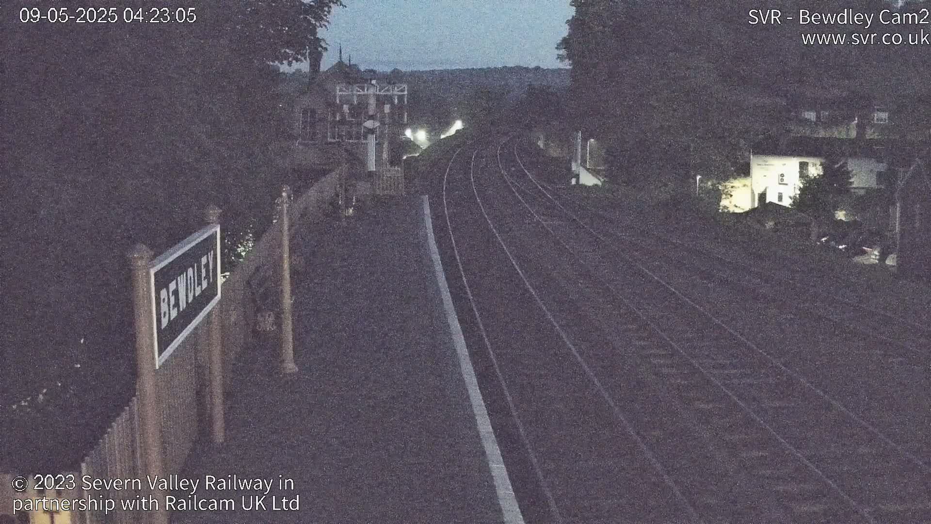 A nighttime view of a train station platform with railway tracks extending into the distance under a dark sky.