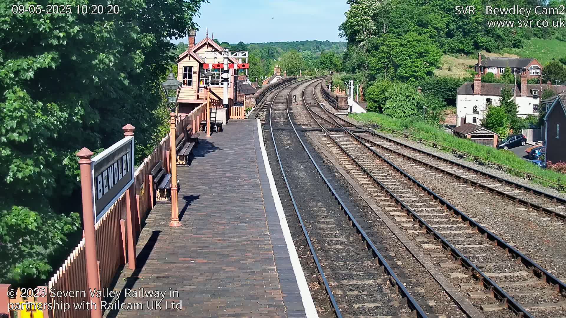 A railway station platform with a signal box and curving tracks extends into the sunny distance, past houses and lush greenery.