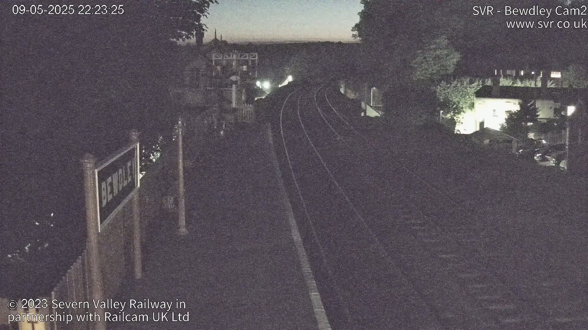 A nighttime view of train tracks at a station, with dimly lit buildings and trees surrounding the area under a dark sky.