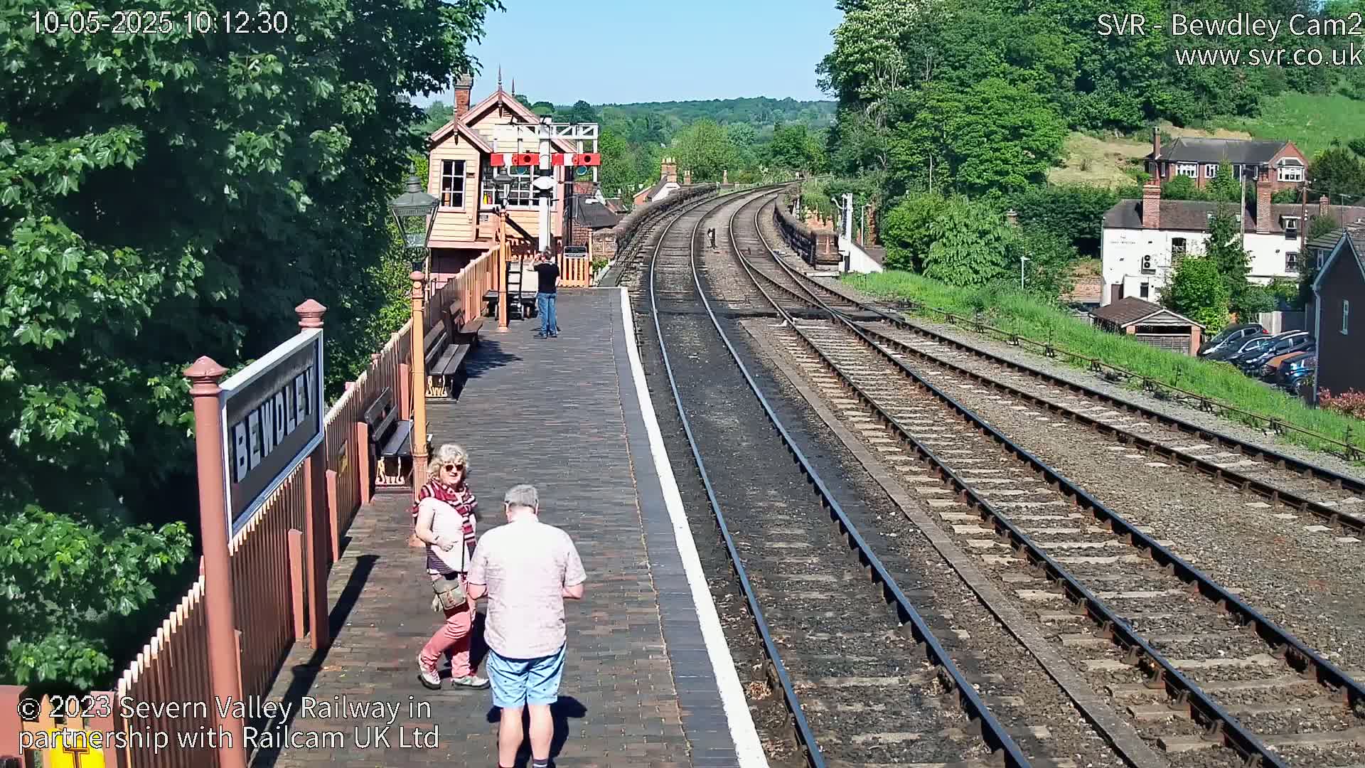 A sunny day at a train station shows a platform with several people, a signal box, and railway tracks curving into the distance.