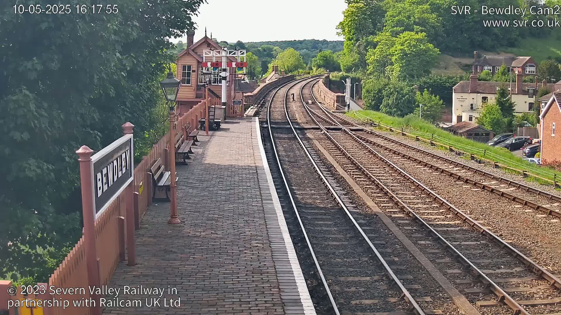 A brick train platform with a signal box overlooks curving train tracks on a sunny day, with lush green trees and residential buildings visible in the background.