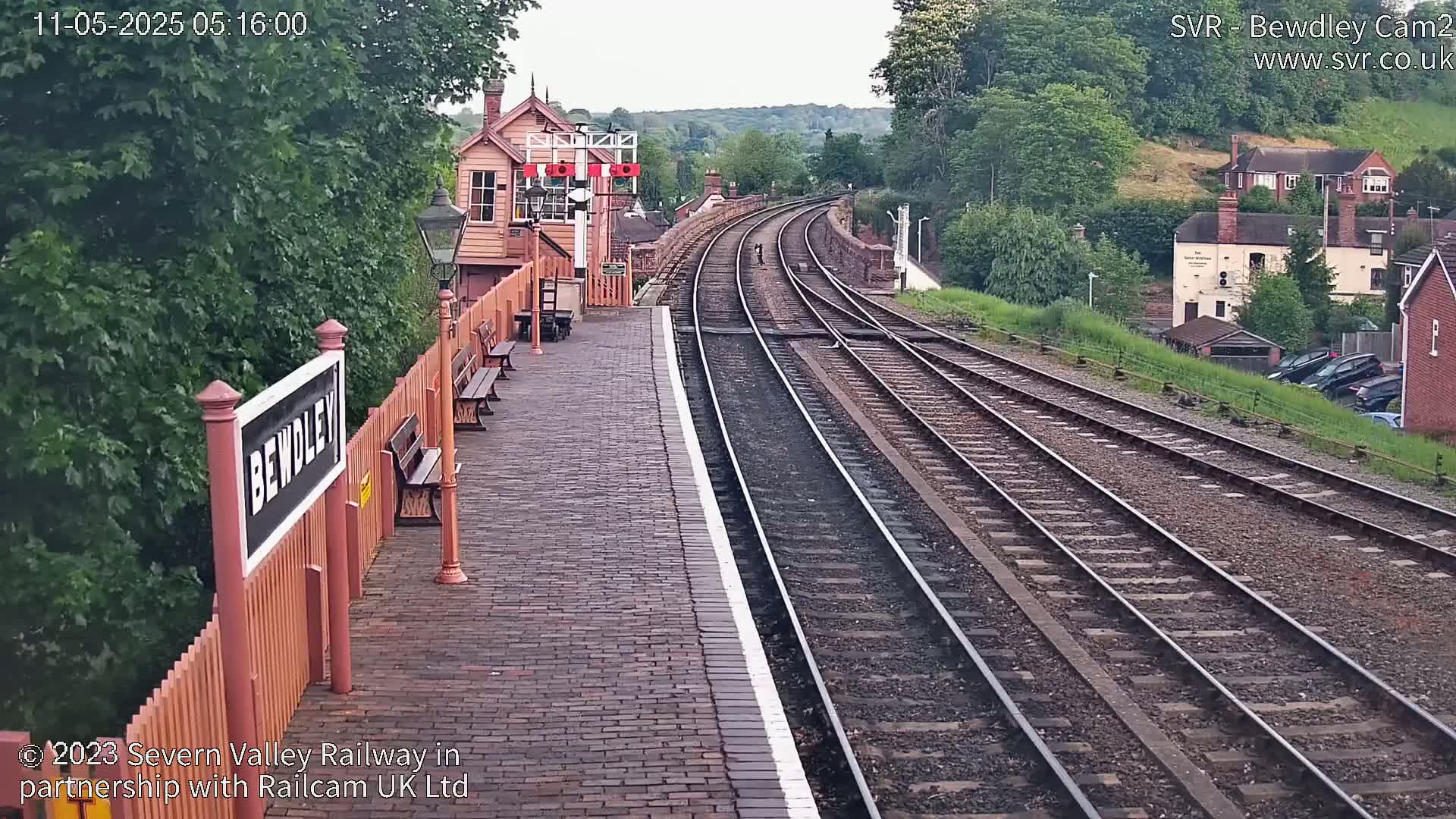A brick railway platform with a signal box and several sets of tracks curves gently away into a tree-lined hillside under an overcast sky.