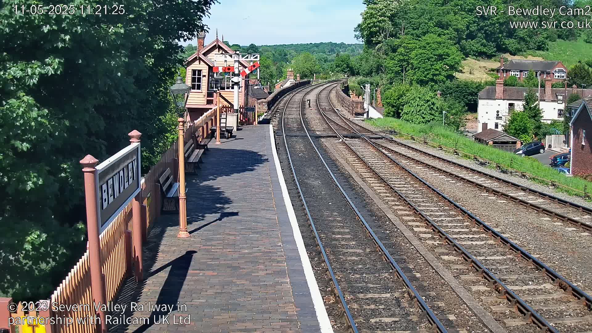 A sunny view of a train station platform with two sets of train tracks curving into the distance, bordered by green hills and buildings.