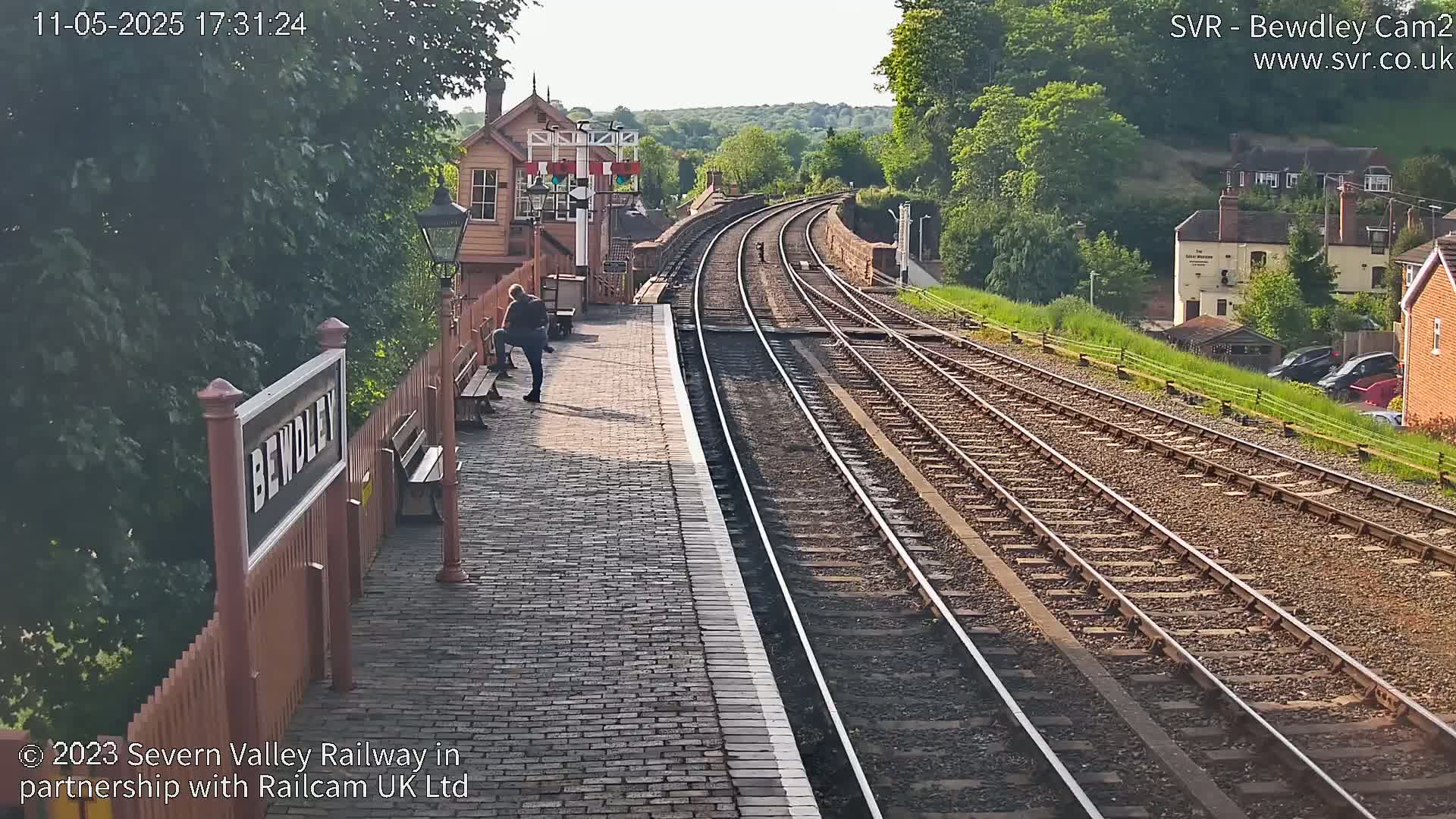 A person sits on a bench at a train station platform next to train tracks extending into the distance on a sunny day.