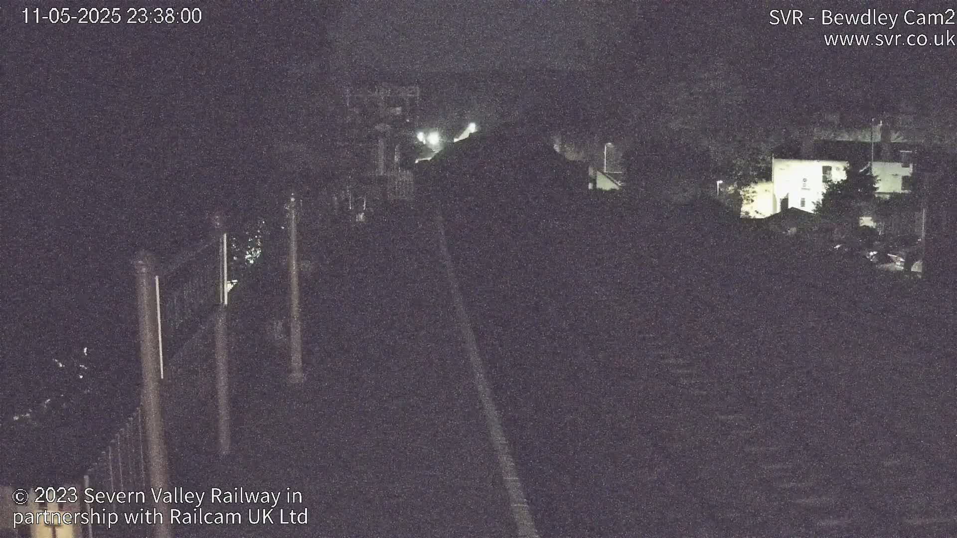 A dark nighttime view of a railway track with dimly lit buildings and trees in the background.