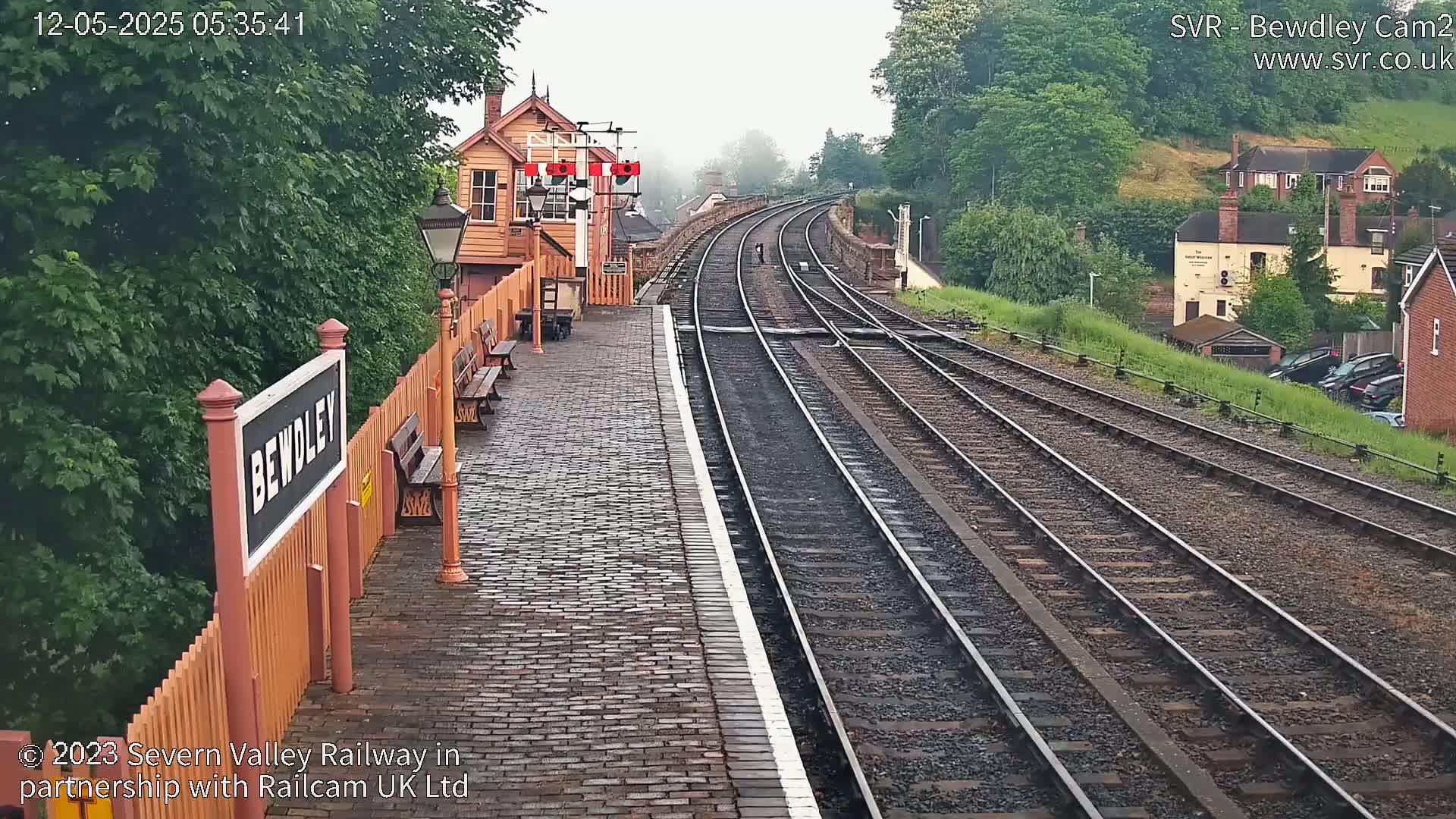 A misty train station platform with a brick walkway next to multiple train tracks, a small wooden building, and a line of trees and houses in the background.