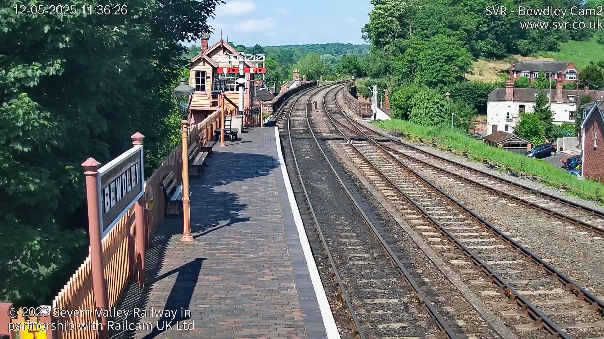 A sunny view of a train station platform with railway tracks curving into the distance, surrounded by lush green trees and buildings.