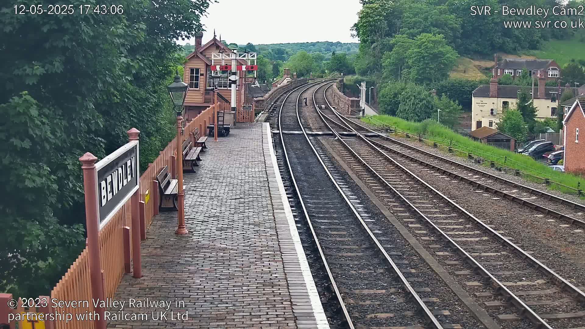 A train platform with a brick surface and wooden benches overlooks curving train tracks that extend into a lush green landscape under an overcast sky.