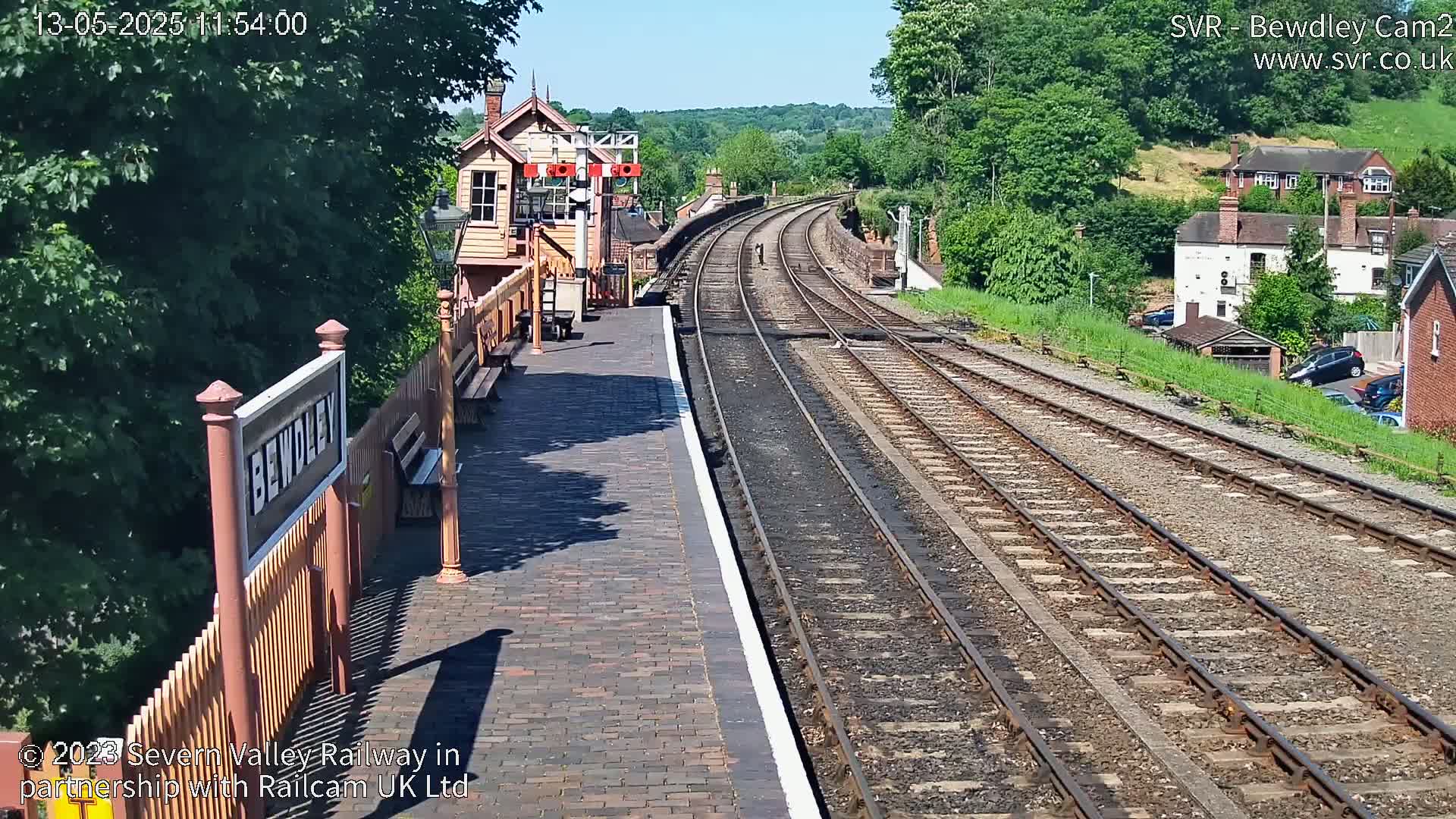 A sunny view of a train station platform with two sets of railway tracks extending into the distance, past a signal box and into a residential area.