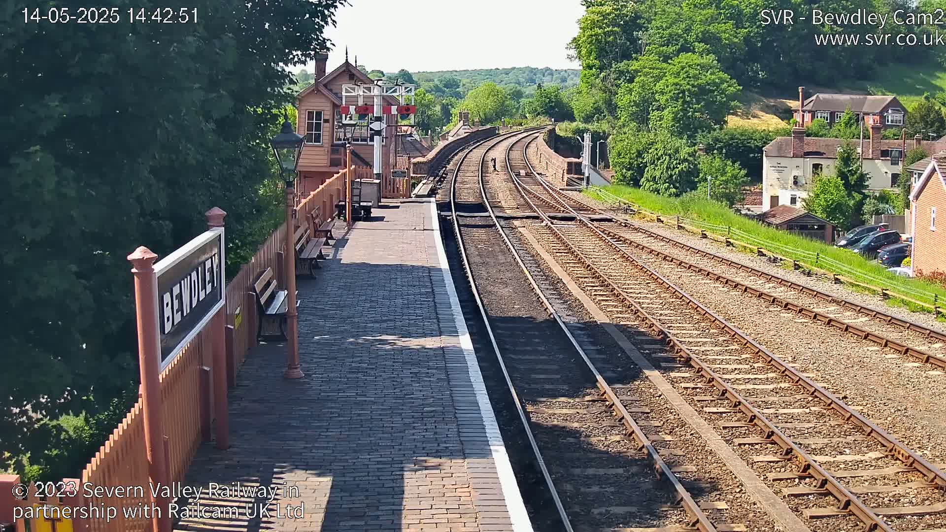 A sunny view of a railway station platform with multiple train tracks extending into the distance, bordered by greenery and buildings.