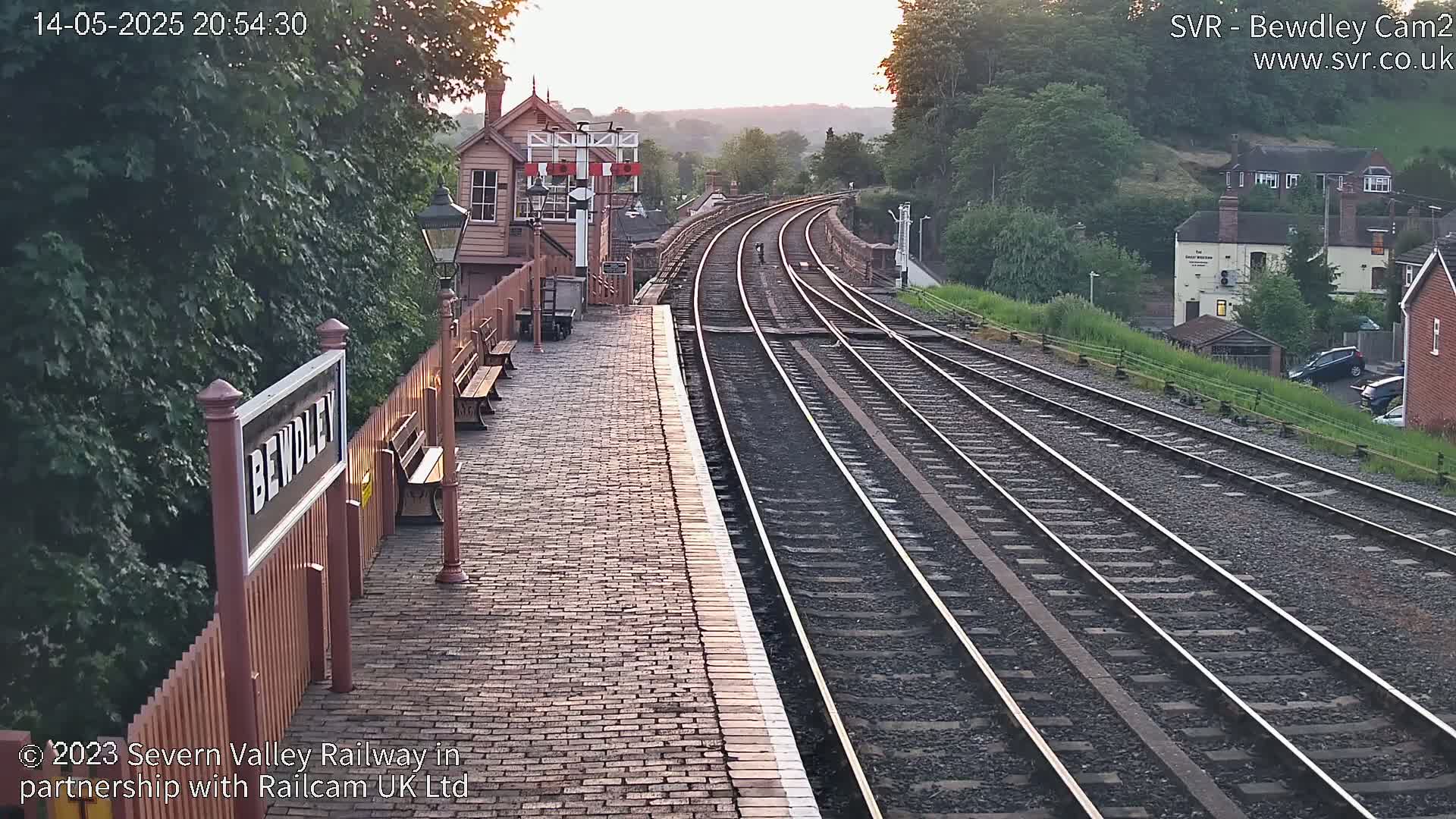 A brick train platform at sunset, with multiple train tracks extending into the distance, bordered by greenery and buildings.