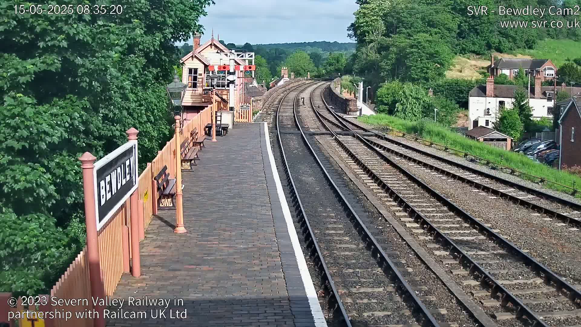 A sunny view of a train station platform with two sets of train tracks curving into the distance, surrounded by green trees and buildings.