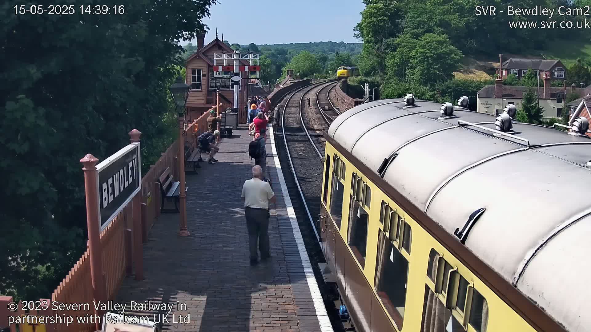 A sunny day at a train station shows a railway platform with people waiting, a train arriving, and another train parked nearby.