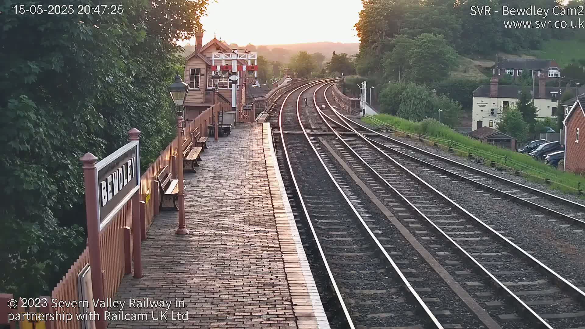 A brick railway platform at sunset, with multiple train tracks extending into the distance, alongside a wooden signal box and a line of parked cars visible beyond a grassy embankment.