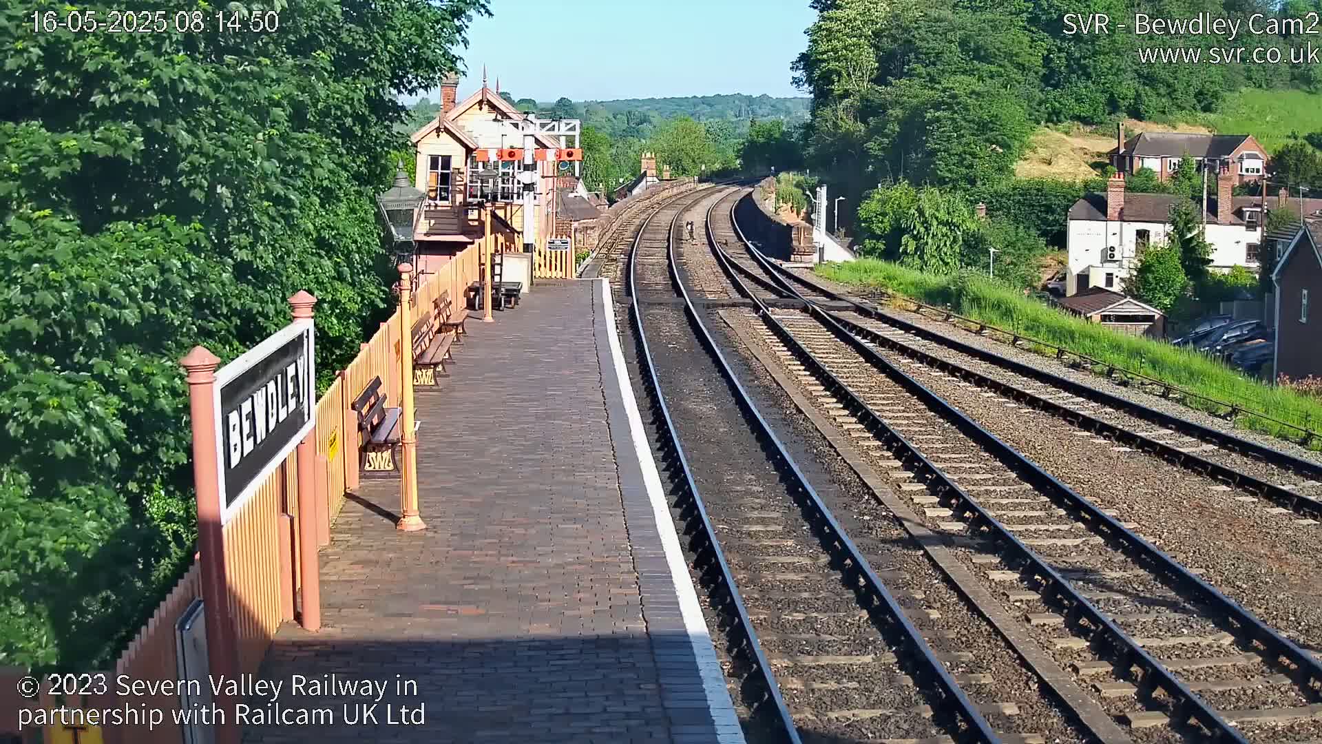 A sunny view of a train station platform with railway tracks extending into the distance, bordered by lush greenery and buildings.