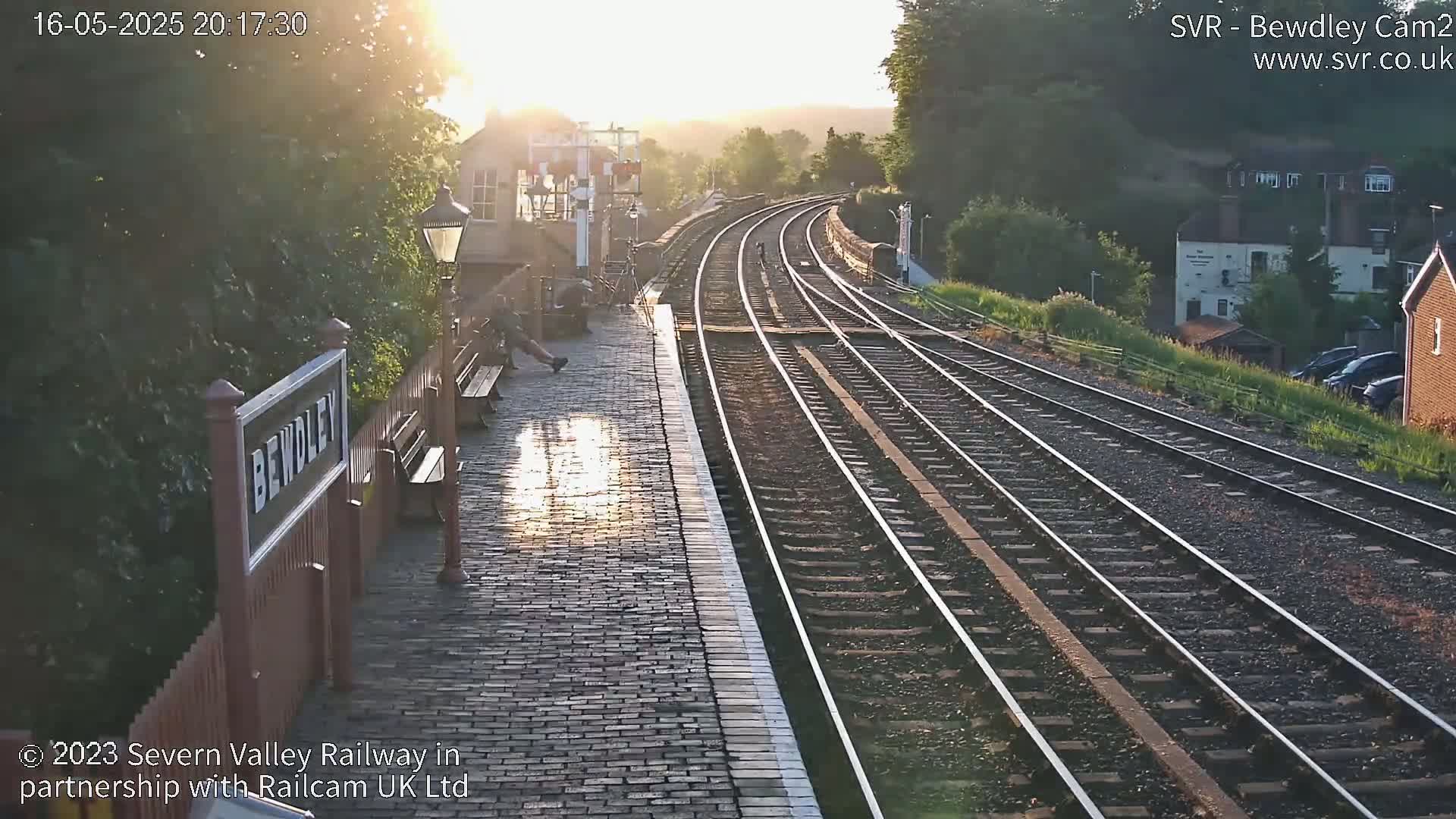 A person sits on a bench at a train station platform next to several sets of train tracks curving into the distance during a sunny evening.