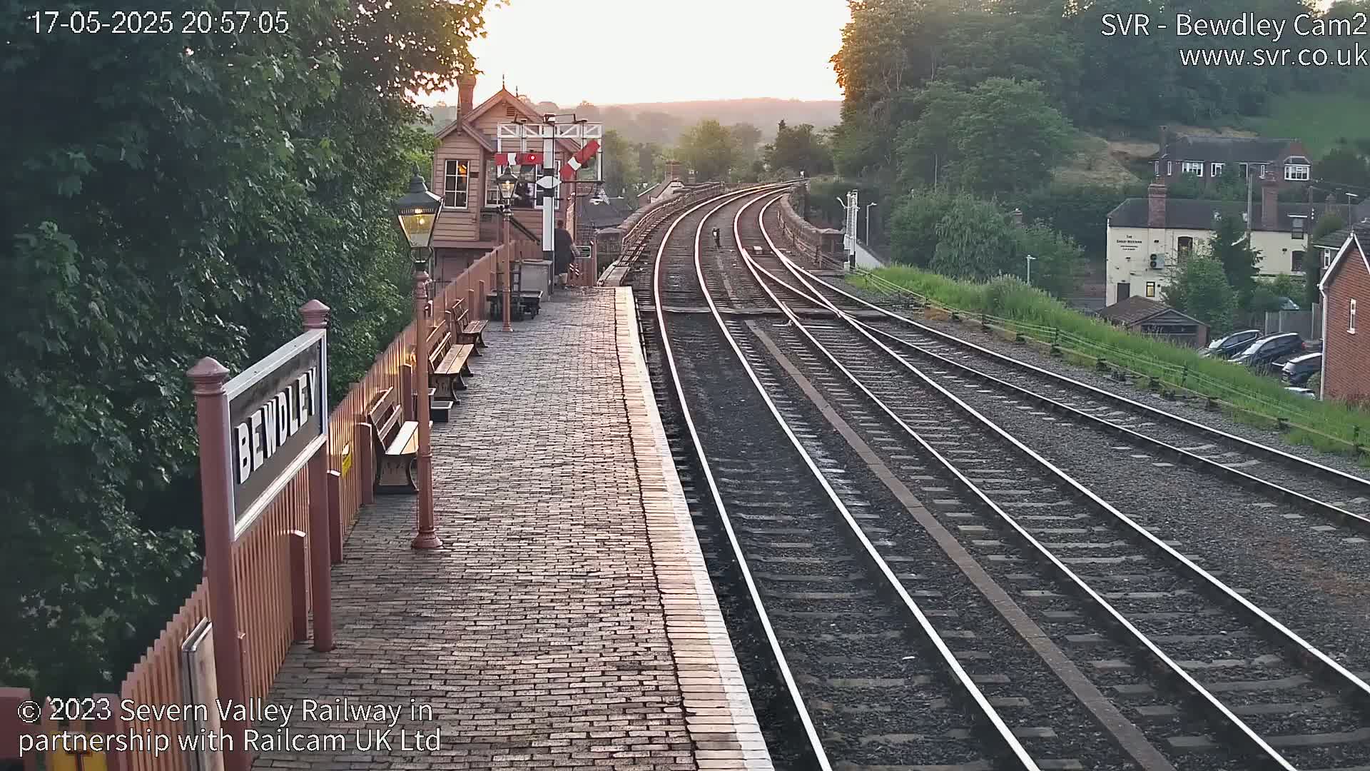 A brick train platform at sunset shows multiple train tracks curving into the distance, bordered by lush green vegetation and a small building.