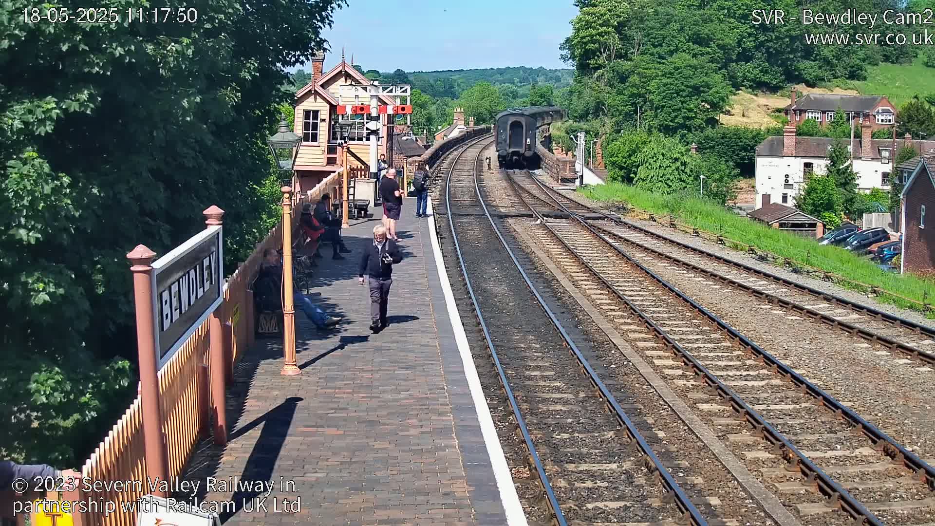 A train is departing a sunny railway station platform where several people are standing and sitting.