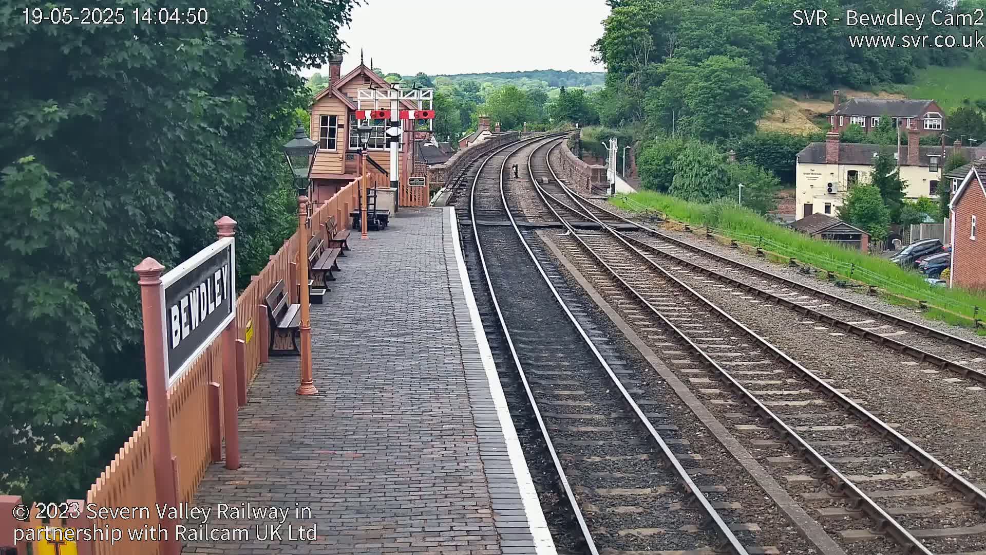 A railway station platform with a brick surface and wooden benches, showing a view of multiple train tracks curving into the distance on a sunny day.