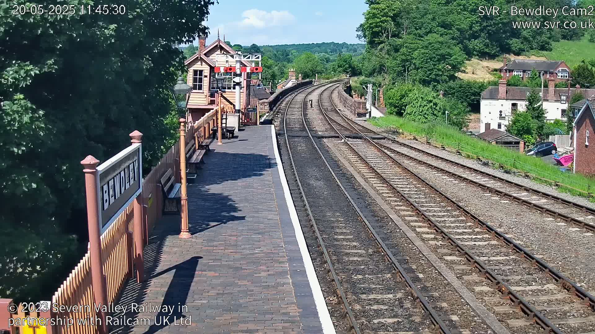 A sunny view of a train station platform with multiple train tracks extending into the distance, surrounded by lush green trees and buildings.