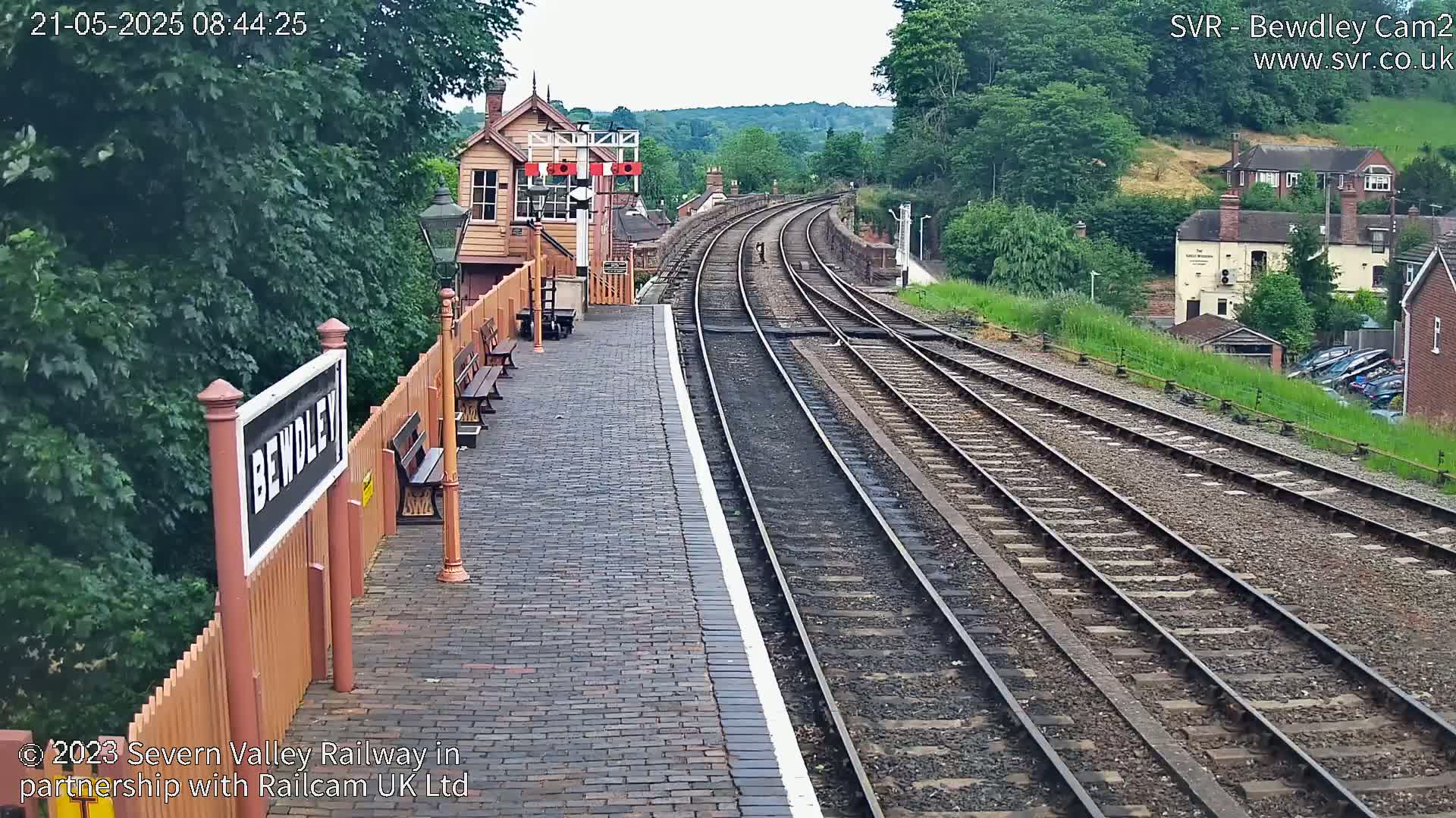 A brick train platform with benches, a signal box, and multiple train tracks curving into a hilly landscape on a partly cloudy day.