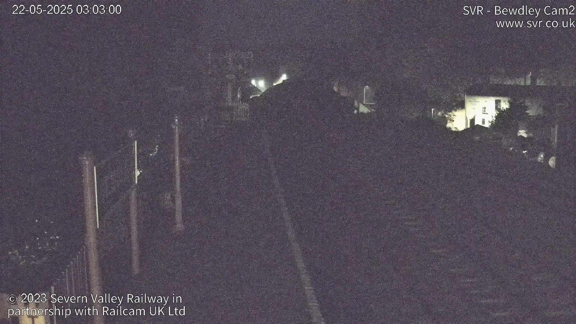 A dark, nighttime view of a railway track, partially obscured by foliage and buildings.