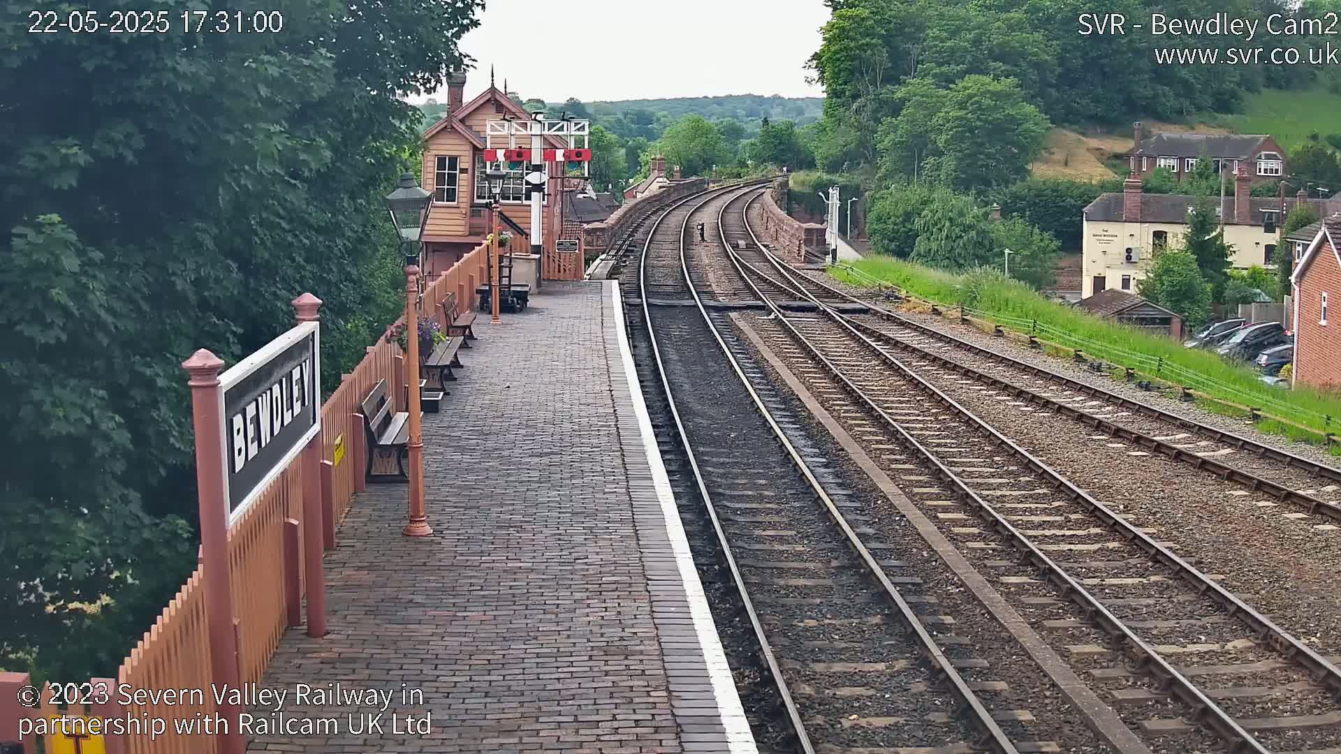 A brick train platform with a wooden sign and signal box, overlooking a pair of train tracks curving into the distance under a partly cloudy sky.