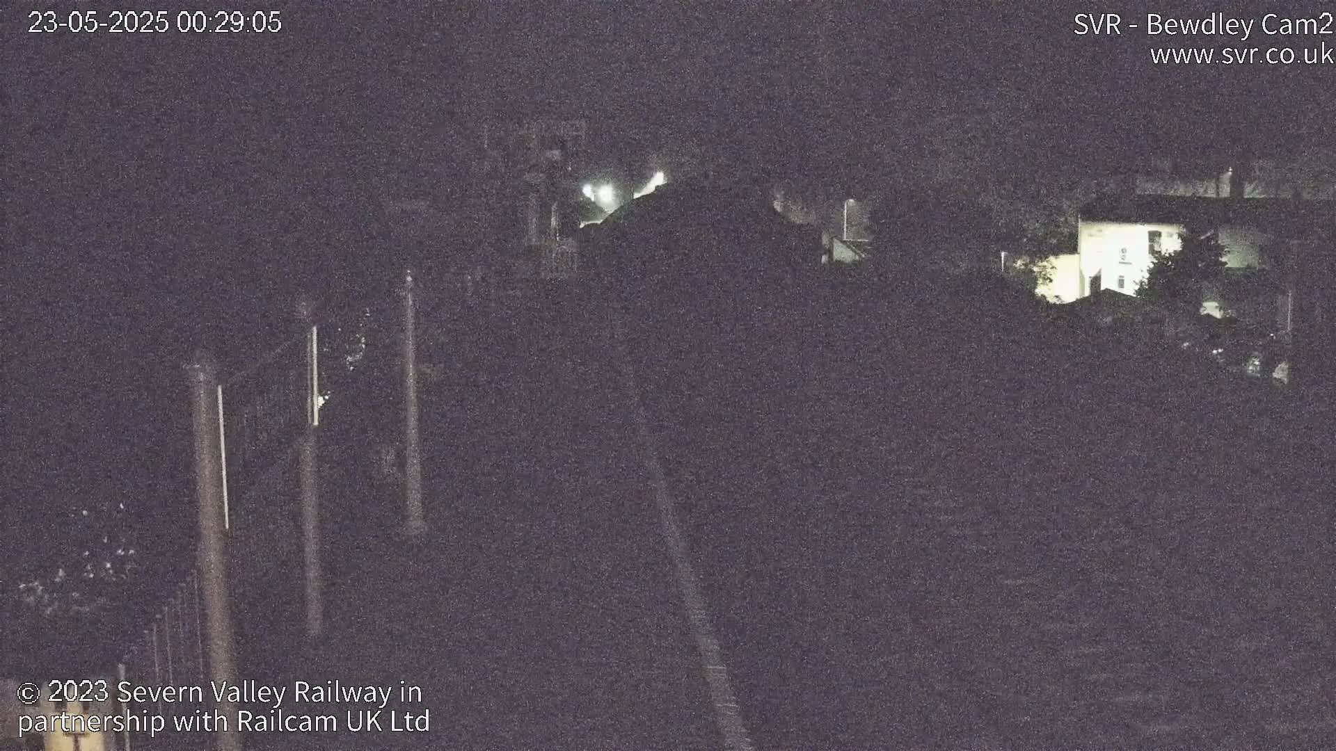 A nighttime, low-light view of a dark, possibly railway, setting with dimly lit buildings and vegetation in the background.