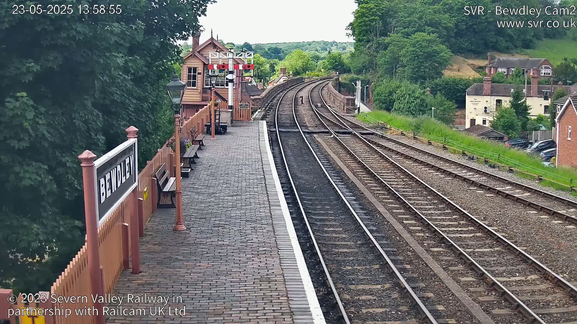 A brick train platform with a wooden sign and a signal box overlooks curving train tracks on a partly sunny day.