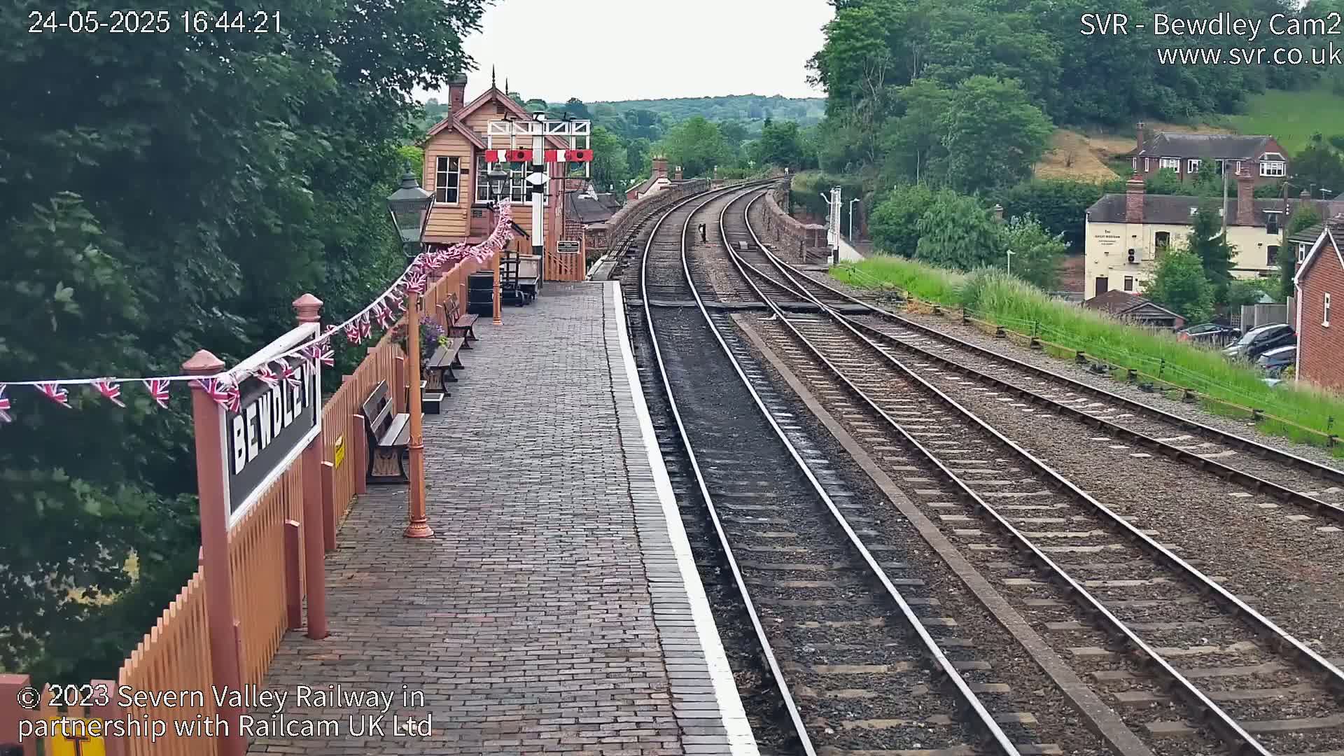 A train station platform with multiple train tracks curves gently upward toward a hillside covered in lush green trees under a partly cloudy sky.