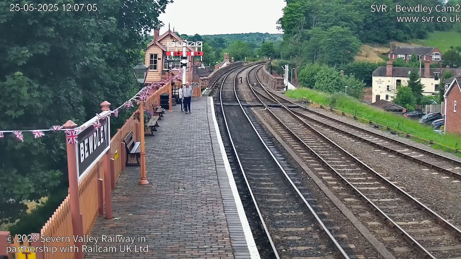 A brick train platform with a signal box and multiple train tracks extends into a slightly overcast, green-tree lined valley.