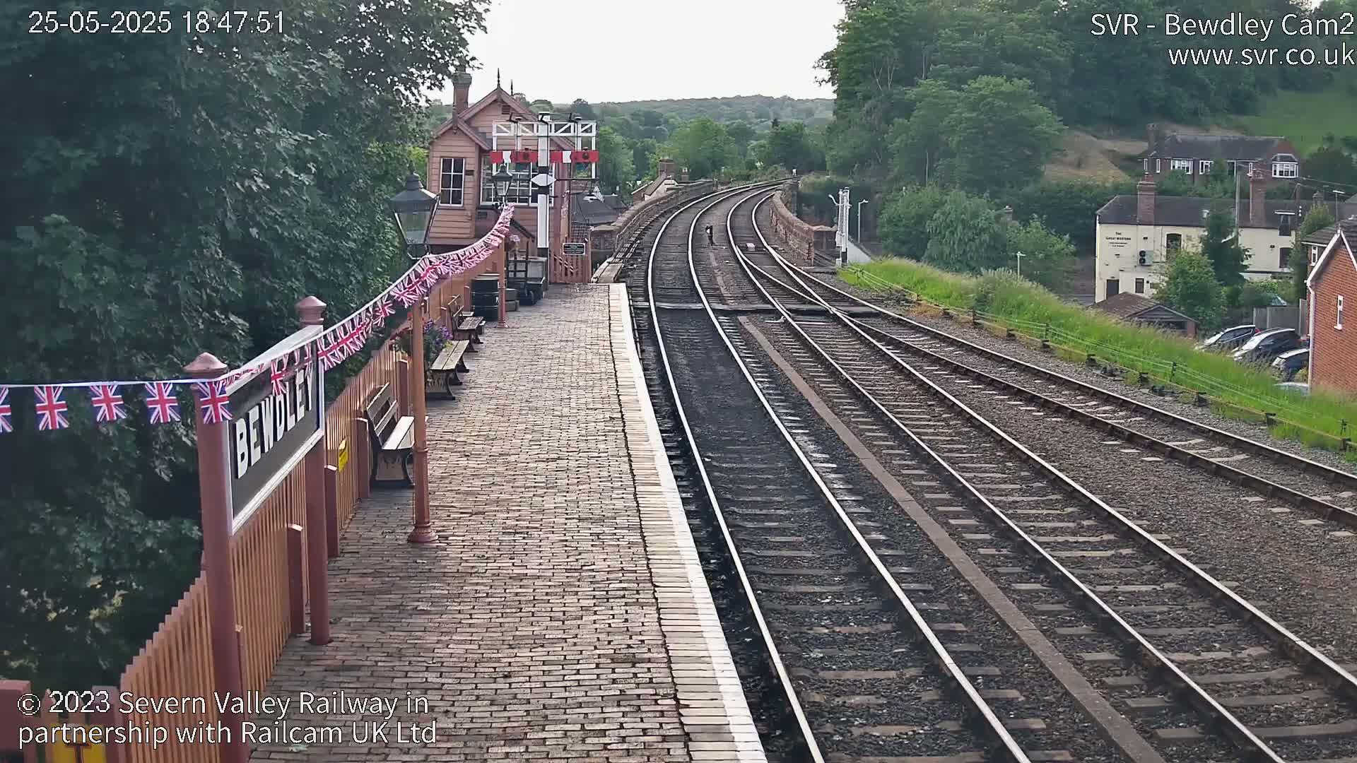 A train station platform with multiple train tracks stretches into the distance under a partly cloudy sky.