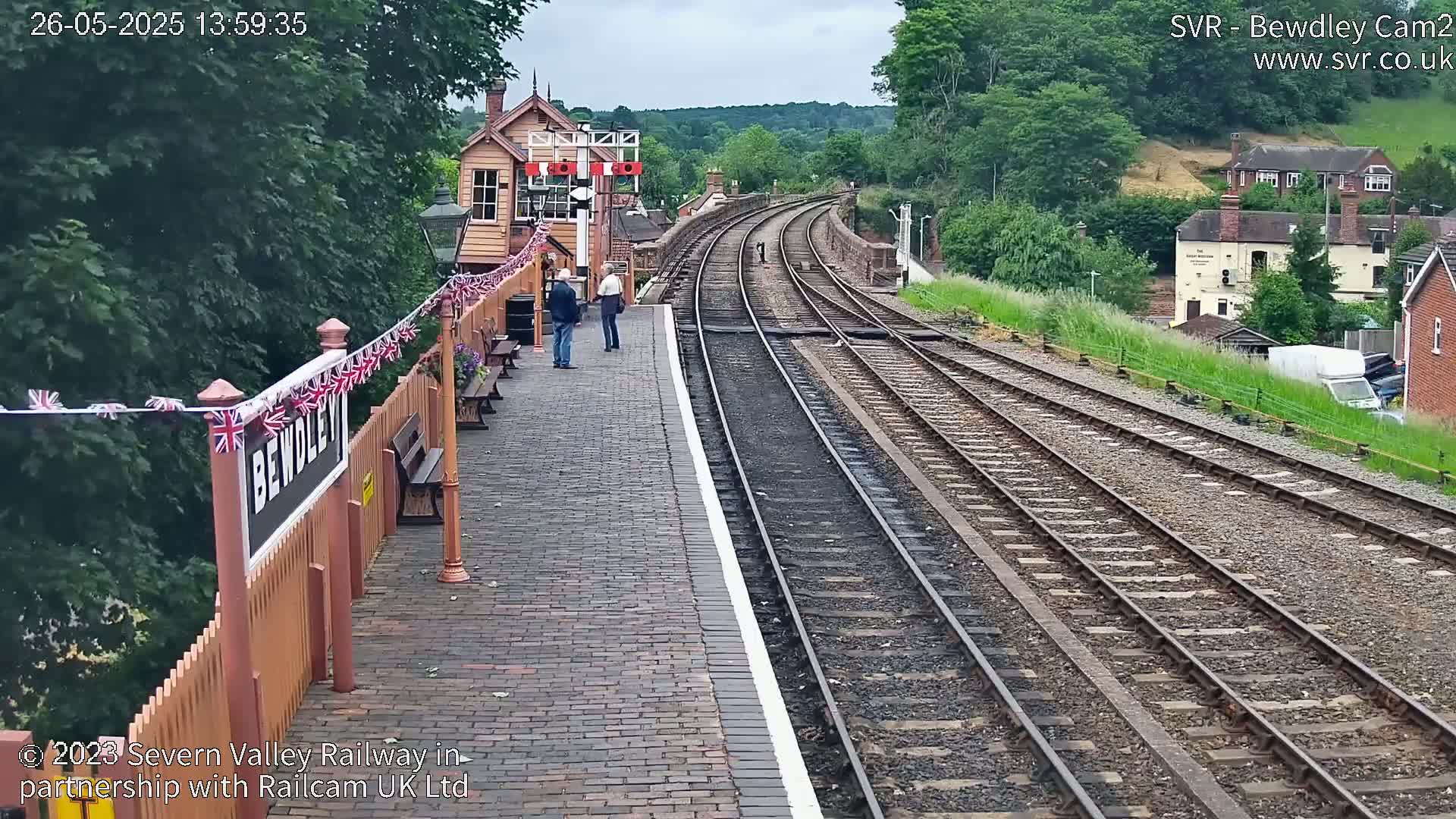 A train station platform with multiple train tracks curving into the distance under an overcast sky, featuring a signal box and a few people standing nearby.