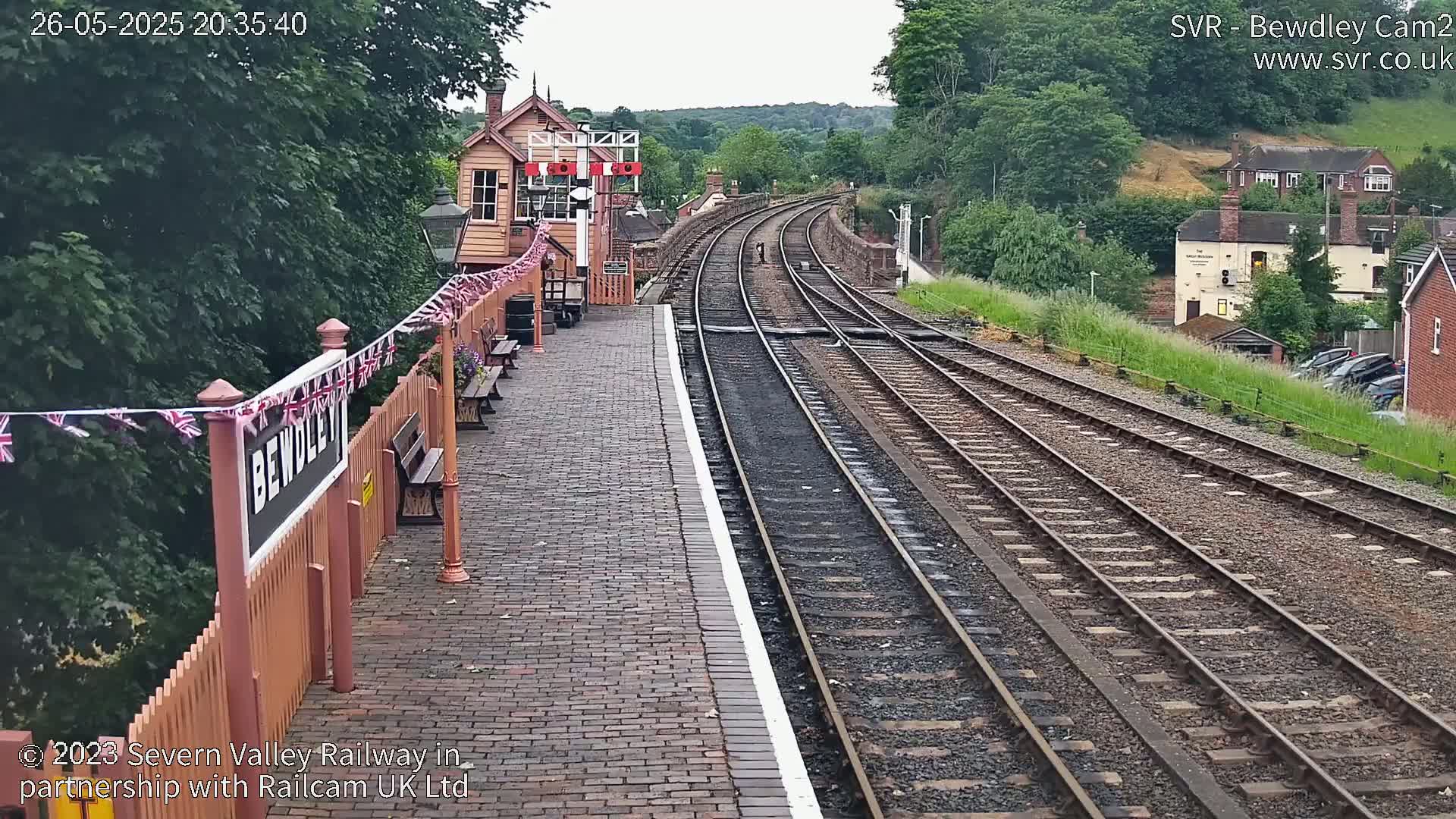A train station platform with a brick pathway, multiple train tracks curving into the distance, and a signal box, all under a partly cloudy sky.