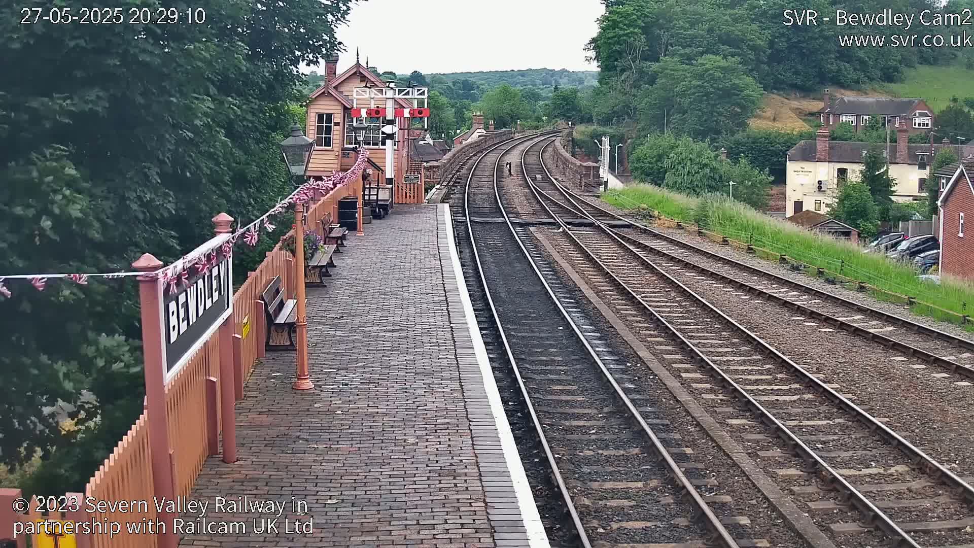 A railway station platform with several tracks extends into the distance under an overcast sky, bordered by lush green vegetation and buildings.