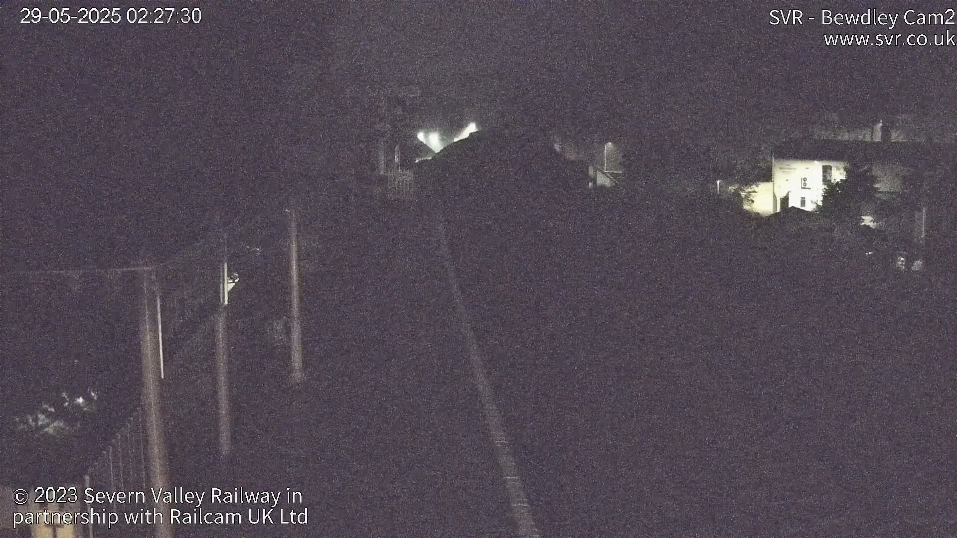 A nighttime, low-light view of a railway track extending into the distance, passing near some buildings and vegetation.