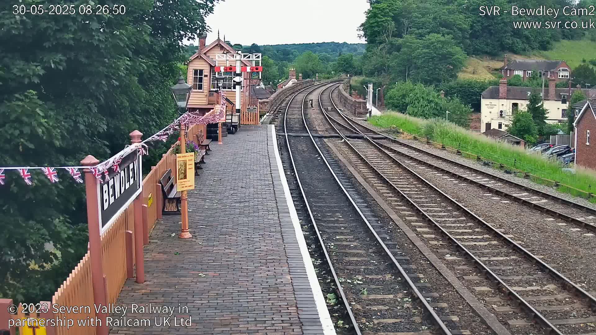 A brick railway platform with a signal box and multiple train tracks extending into the distance on a partly cloudy day.