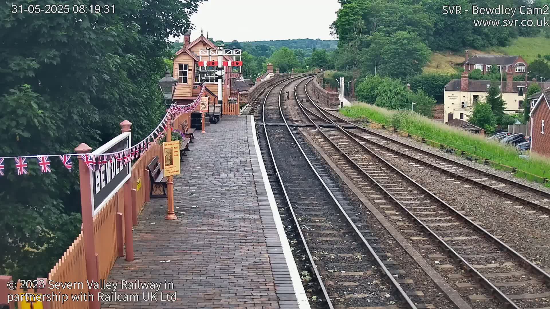 A train station platform with brick paving and a wooden fence decorated with British flags overlooks curving train tracks extending into a green hillside under a partly cloudy sky.