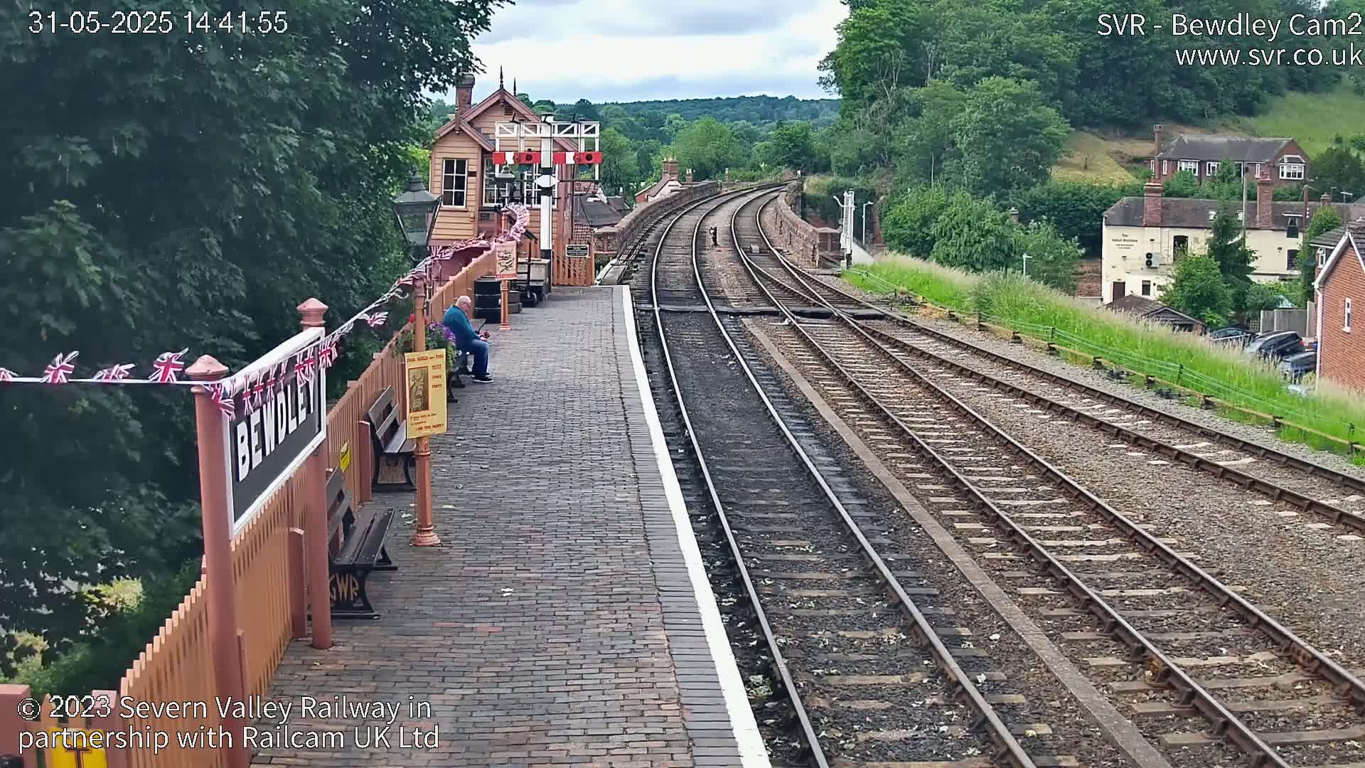 A brick train platform with a man sitting on a bench overlooks a pair of train tracks curving into a verdant hillside on a partly cloudy day.