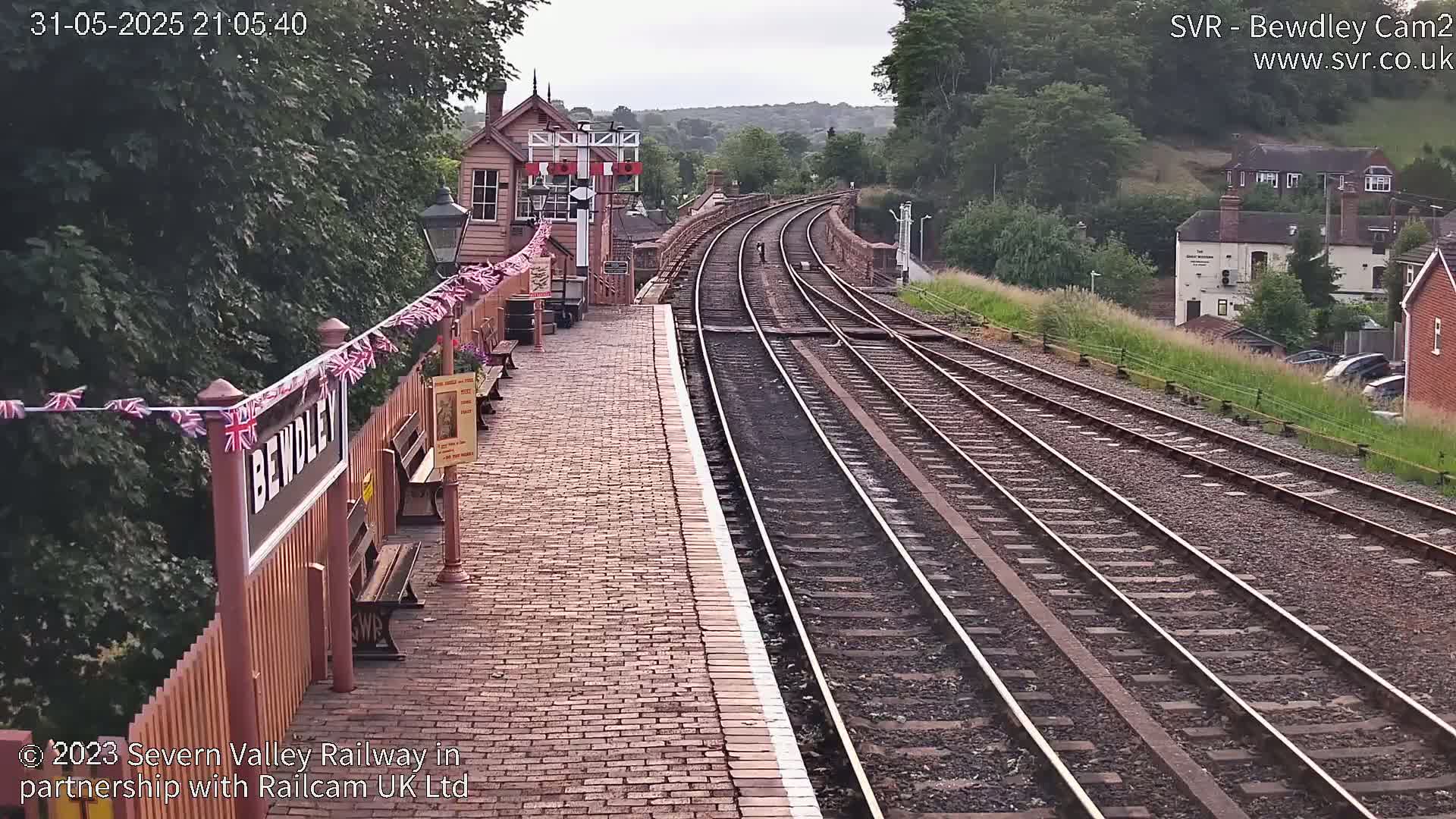 A brick train platform with a station building and signal box, decorated with bunting, overlooks a curving railway track extending into a hilly, wooded area under a partly cloudy sky.