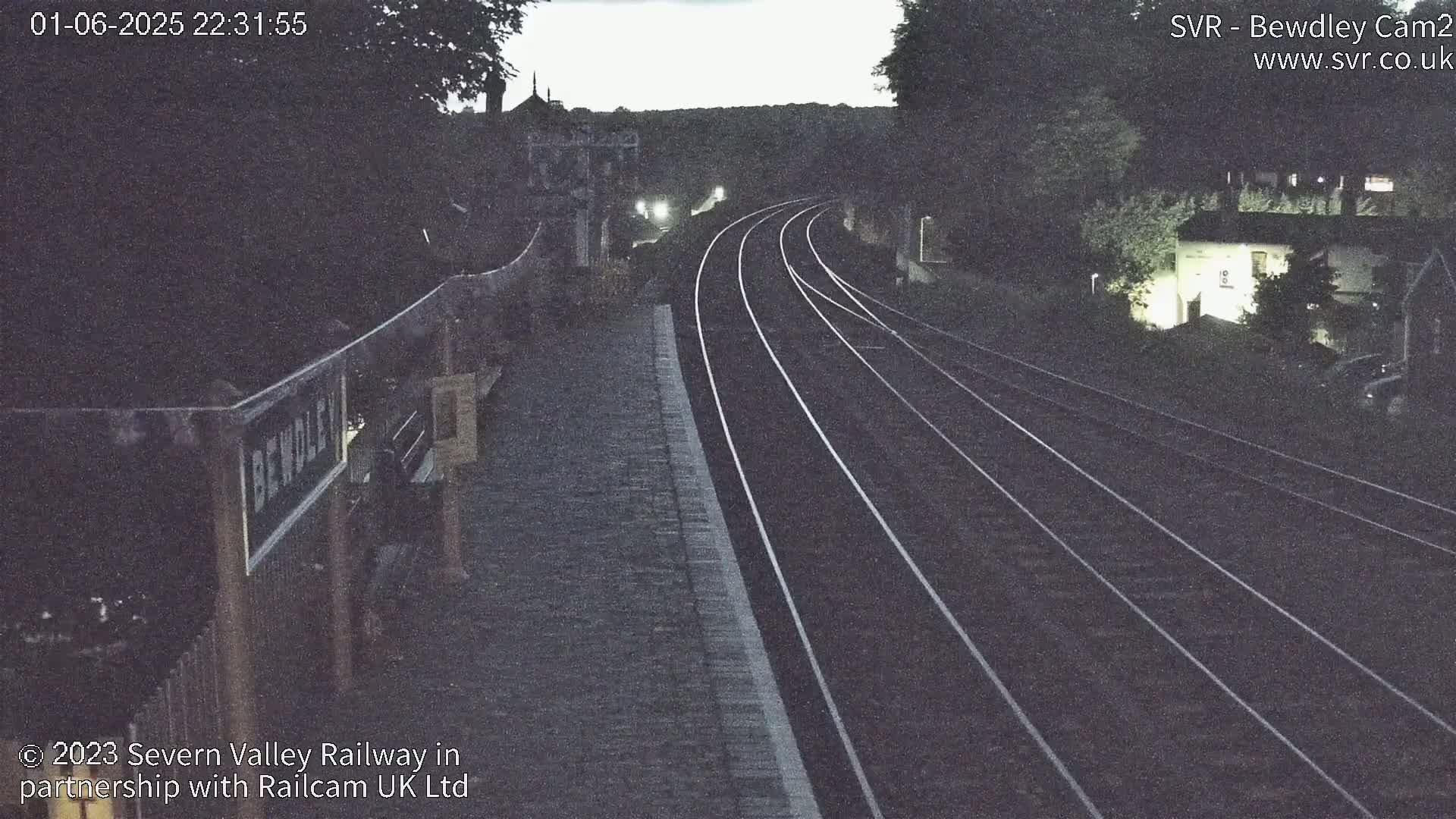 A nighttime view of a train station platform with railway tracks curving into the distance under a dark sky.
