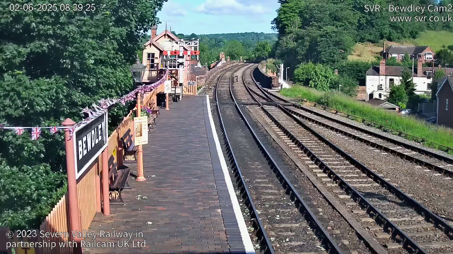 A sunny view of a railway station platform and tracks, with lush green trees and buildings in the background.
