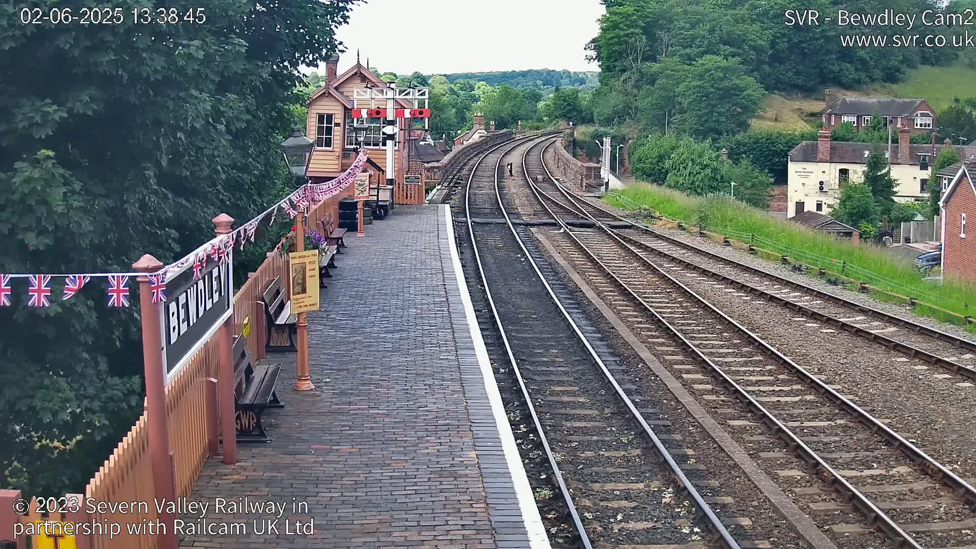 A train station platform with brick paving and two sets of train tracks extending into the distance on an overcast day, with a signal box and bunting visible.
