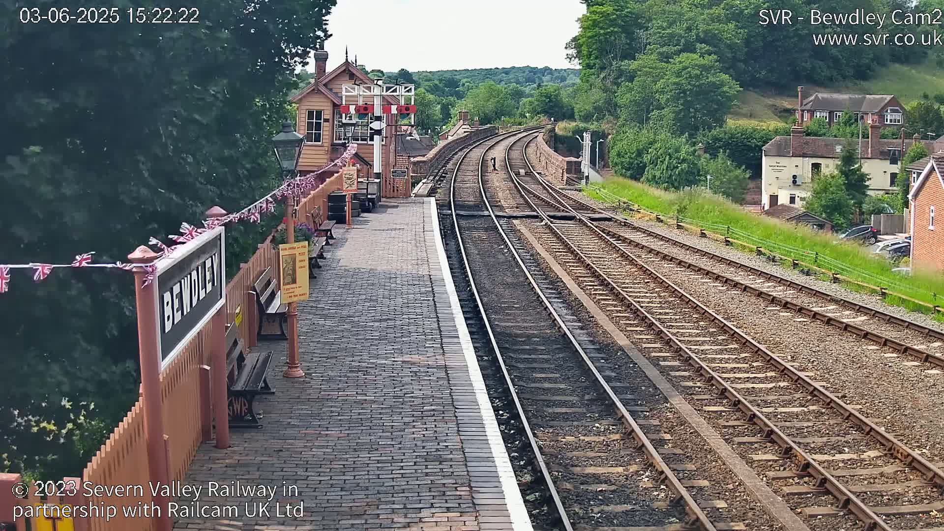 A brick railway platform with a wooden station building and curving train tracks extends into a sunny, green hillside.