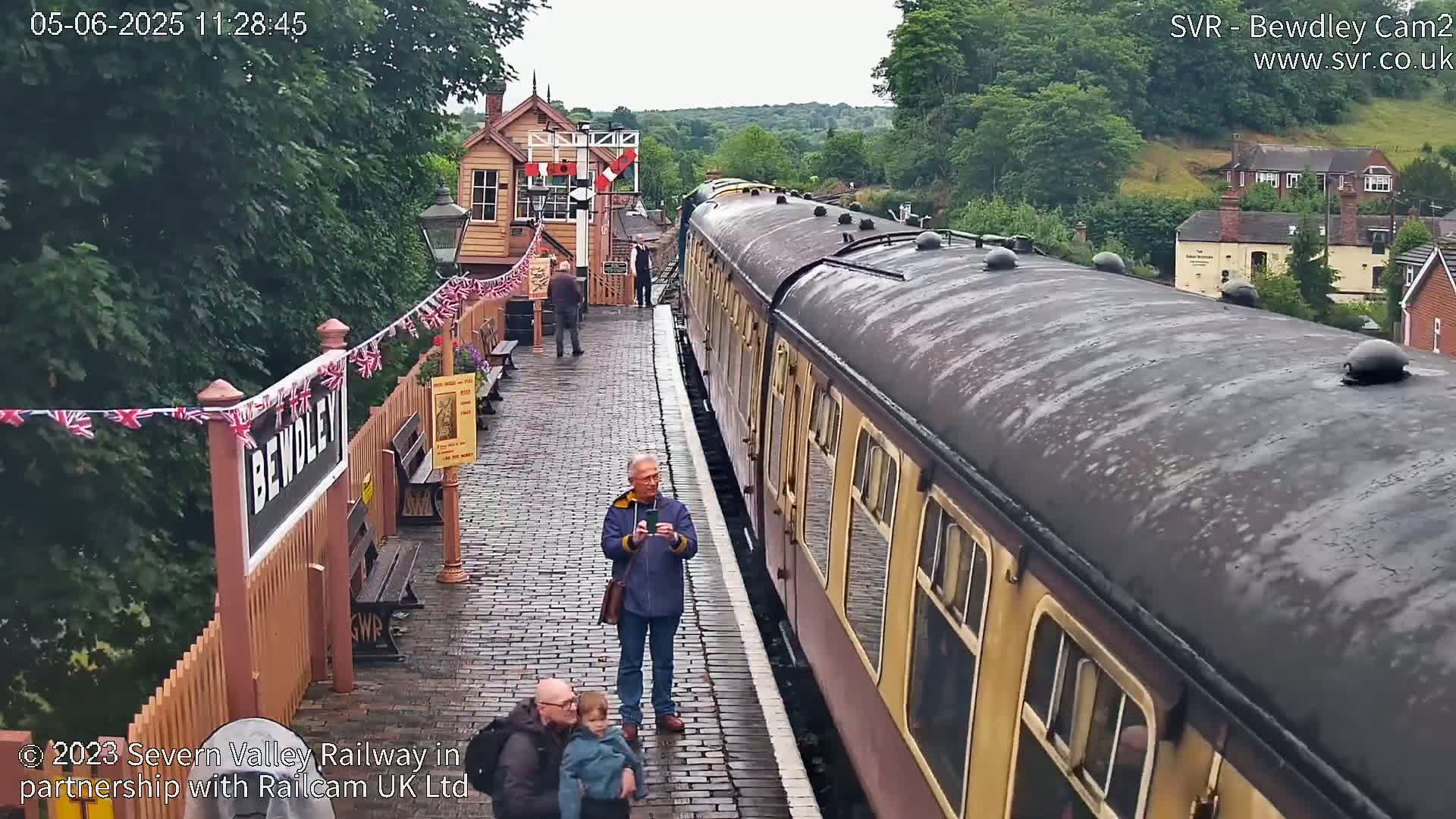 A long passenger train is at a station on a wet, overcast day; several people stand on the platform, some taking photographs.
