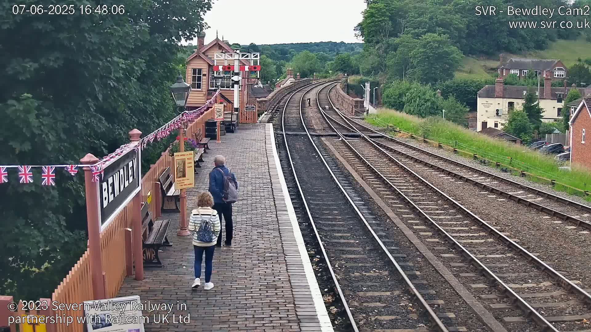 A train station platform with two people walking away from the camera, multiple train tracks curving into the distance, and a partly cloudy sky.